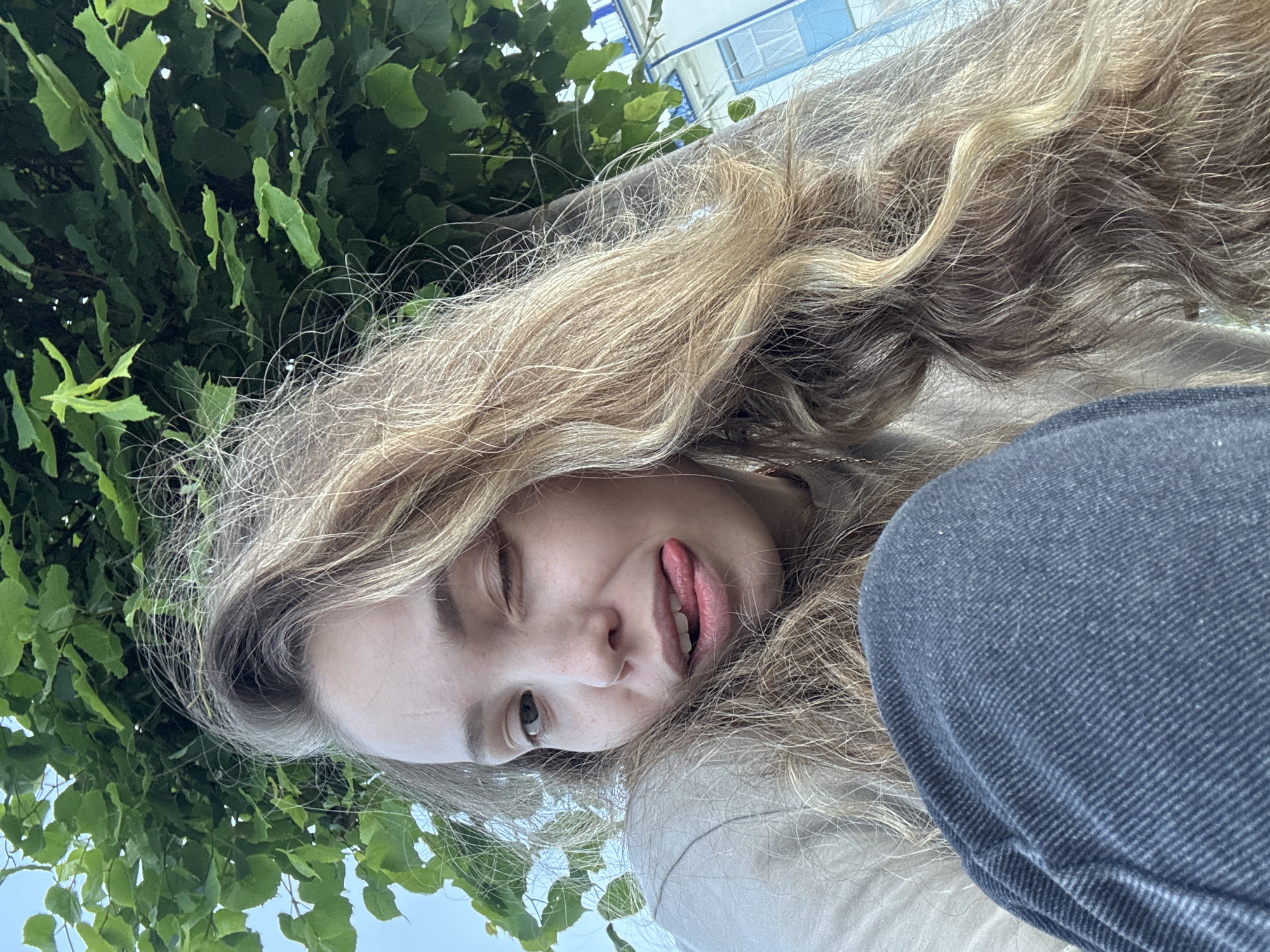 A young Caucasian woman with long, wavy blonde hair is pictured outdoors, likely in a park or near a body of water, given the lush green foliage and the glimpse of a building with blue architectural details in the background. She is wearing a neutral-colored long-sleeved shirt and dark gray pants. The woman is looking directly at the camera, winking and sticking out her tongue playfully. Her facial features are visible, including freckles on her nose and cheeks, and a faint scar on her forehead. The lighting suggests it is daytime. The background includes green leaves from a tree overhead and a building with white walls and blue windows, possibly in the city of Chaikovsky, Russia.