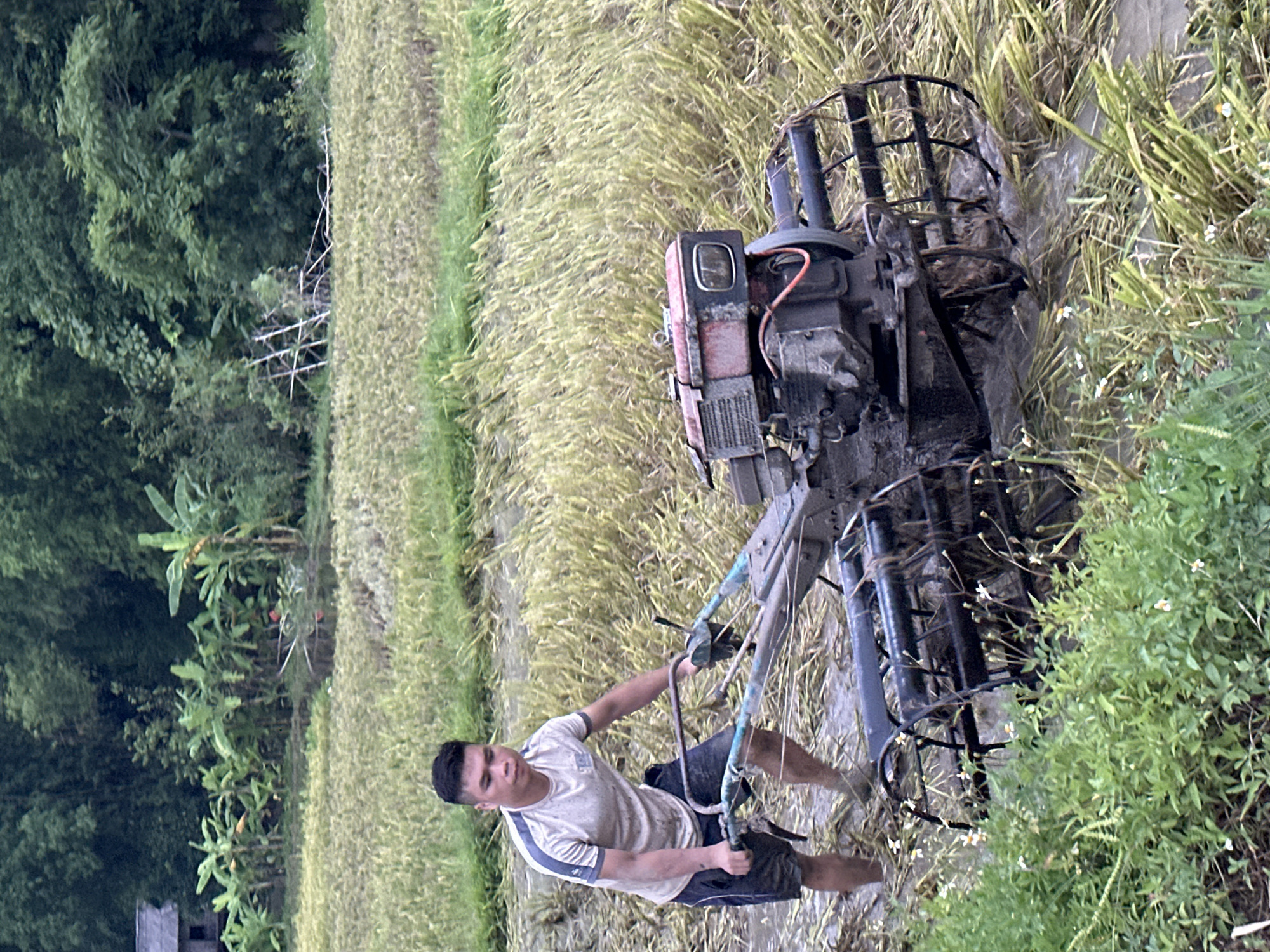 A young man in a light-colored t-shirt and dark shorts is operating a walking tractor in a muddy rice field in Xóm Khán, Vietnam. The field is filled with stubble from a harvested crop and water, indicated by the splashing around the tractor's tines. The man is holding onto the handlebars of the tractor, appearing to guide it through the mud. The background shows lush green trees and vegetation, with a small building visible on the left side of the frame. The overall scene depicts agricultural work in a rural Vietnamese setting.