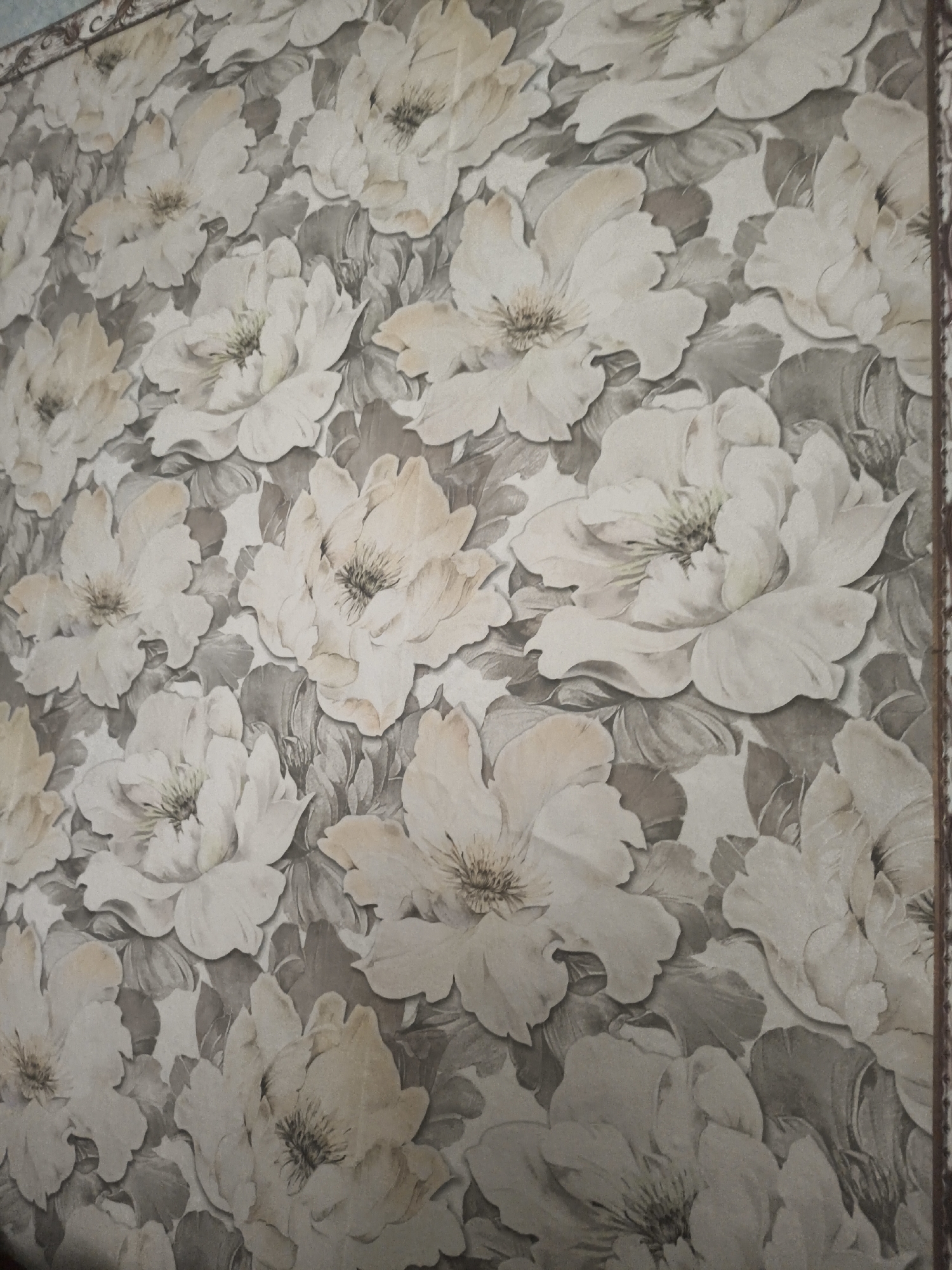 A close-up, head-on shot shows a wallpaper pattern featuring large, detailed floral designs with subtle hints of pale peach and cream on a muted gray background. The flowers are rendered with a sense of depth and texture, with visible stamens and layered petals. The image is a still life, devoid of any human presence or urban context within the city of Умыган, Russia.