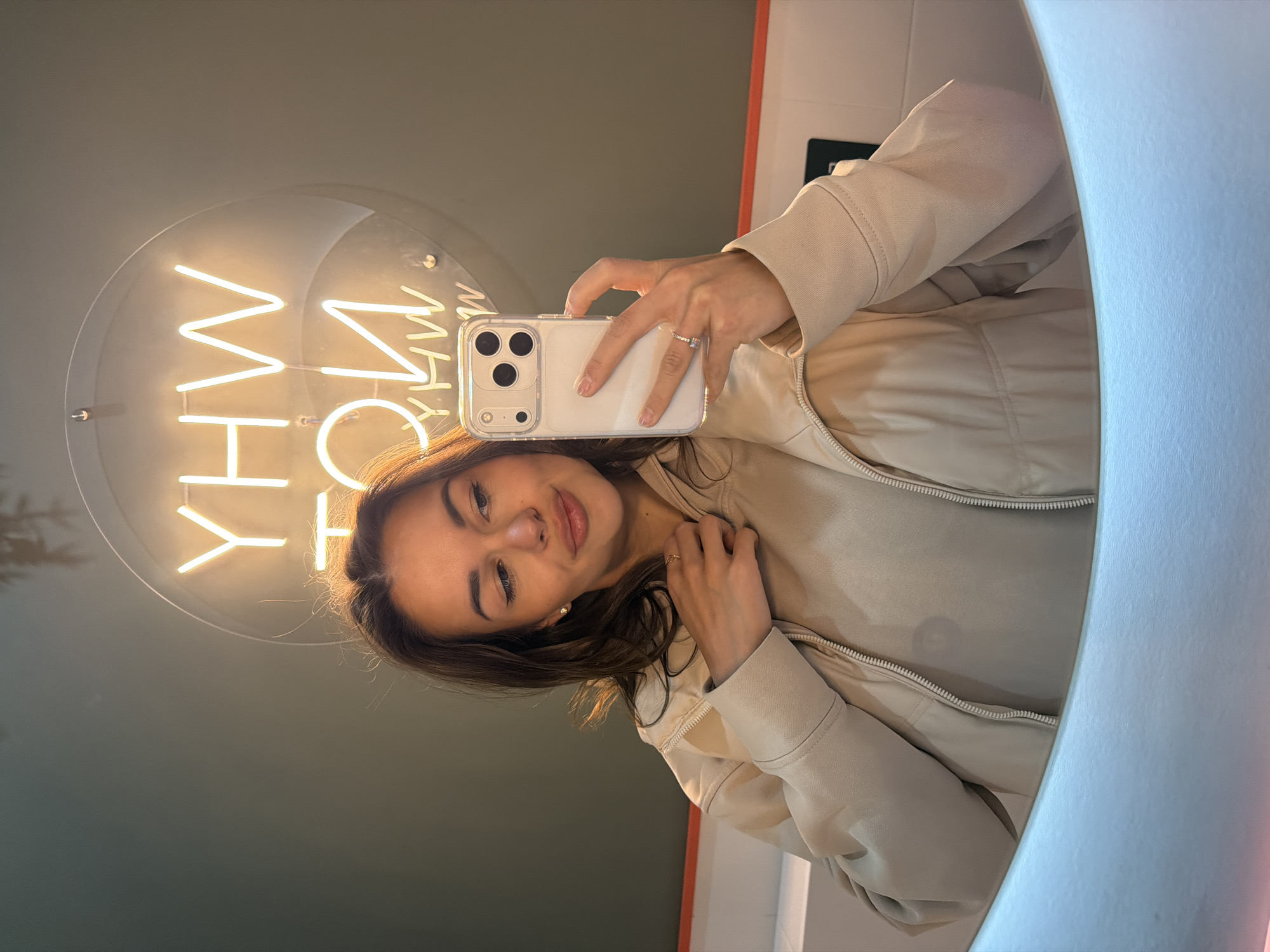 A woman is taking a selfie in a mirror. She is wearing a light-colored jacket over a light-colored shirt and holding a white iPhone 13 or 14 with a clear case. The background includes a neon sign that reads "WHY" vertically. The location is potentially in Krasnoyarsk, Russia.