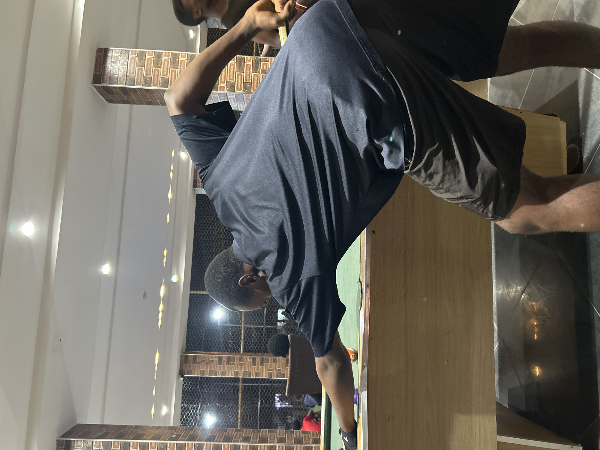 A man in a dark t-shirt and shorts is playing billiards in what appears to be a dimly lit indoor entertainment venue in Ihiagwa, Nigeria. He is leaning over the green billiard table, cueing a ball. Two billiard balls, one orange and one purple, are visible on the table. The background shows a brick-patterned wall, a caged window, and other patrons in the distance. The lighting appears to be artificial, with ceiling lights casting a warm glow. The floor is tiled.