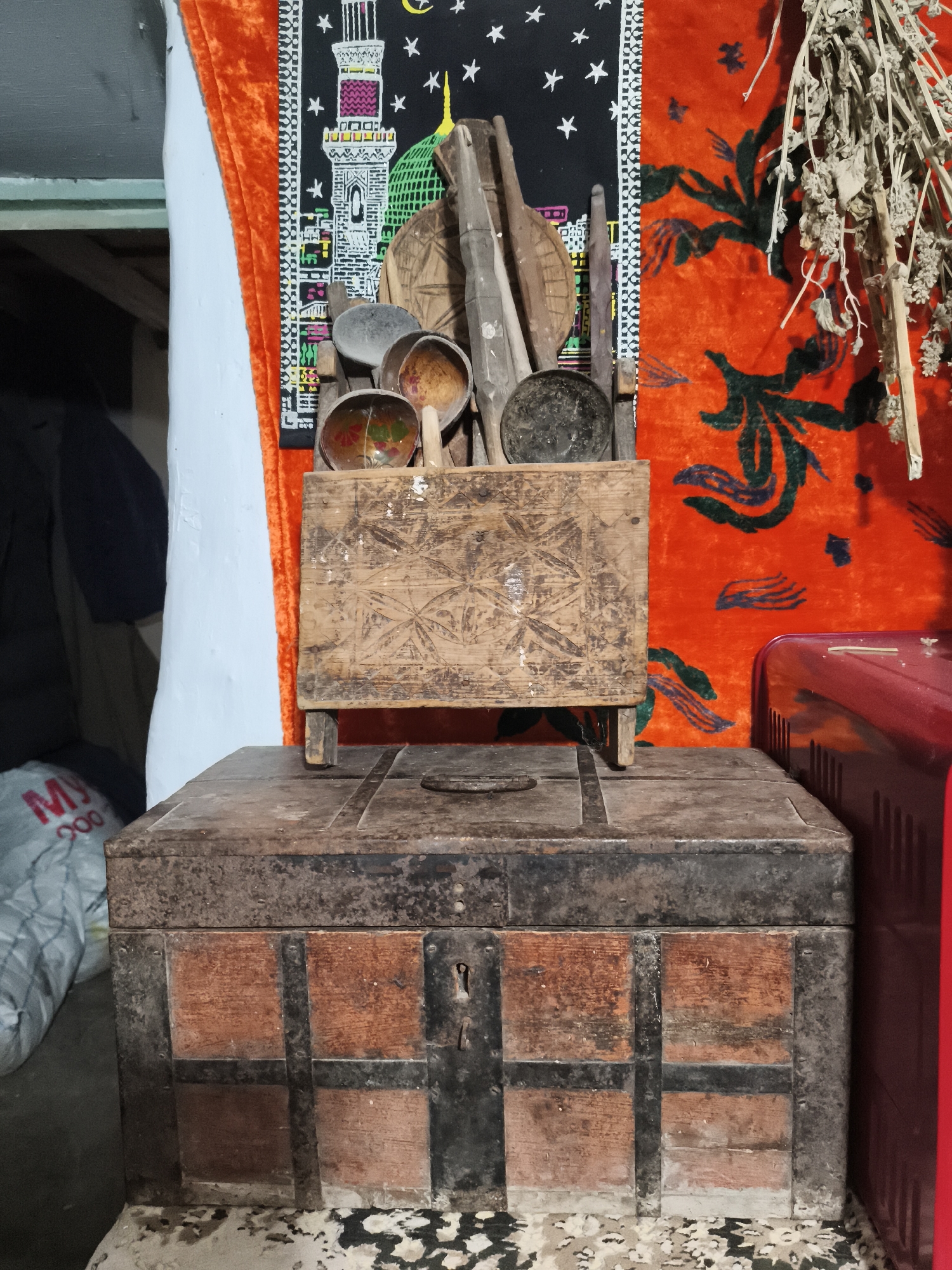 The image depicts an interior scene, possibly a home or storage area, with a collection of old, rustic objects. In the foreground, a dark, metal-bound wooden chest sits on a surface with a patterned, speckled finish. The chest has visible metal bands and a keyhole. On top of the chest, there is a smaller, intricately carved wooden box with a patterned lid. Behind this, various wooden utensils and bowls are arranged. The background features vibrant, patterned fabric, one piece a deep orange with a dark green floral motif, and another a black fabric with white stars and an illustration of a mosque. To the right, a portion of a red, ribbed object, possibly a metal appliance or furniture, is visible. The lighting is dim, suggesting an indoor setting, and there are no people present in the frame. The overall impression is one of aged items and a lived-in space.