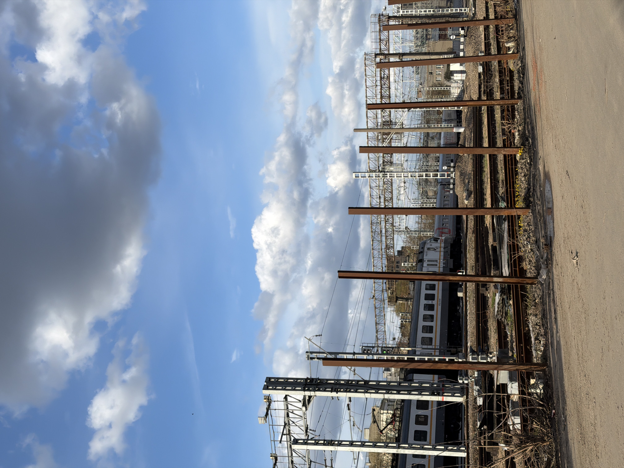 The image depicts a view of railroad tracks and a train in Moscow, Russia. The sky is partially cloudy. A series of rusty metal posts and overhead power lines obscure the view of the train and the railway infrastructure. To the right of the tracks, there is a concrete or asphalt surface.