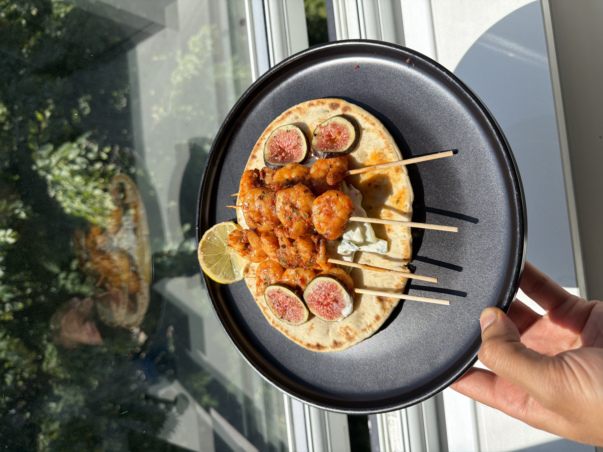 A person's hand holds a black plate containing a meal of shrimp skewers on flatbread, garnished with figs and a lemon slice. The meal is illuminated by natural sunlight, casting shadows on a white surface below. In the background, a reflection in glass shows greenery and an outdoor scene. The location is implied to be in London, United Kingdom.