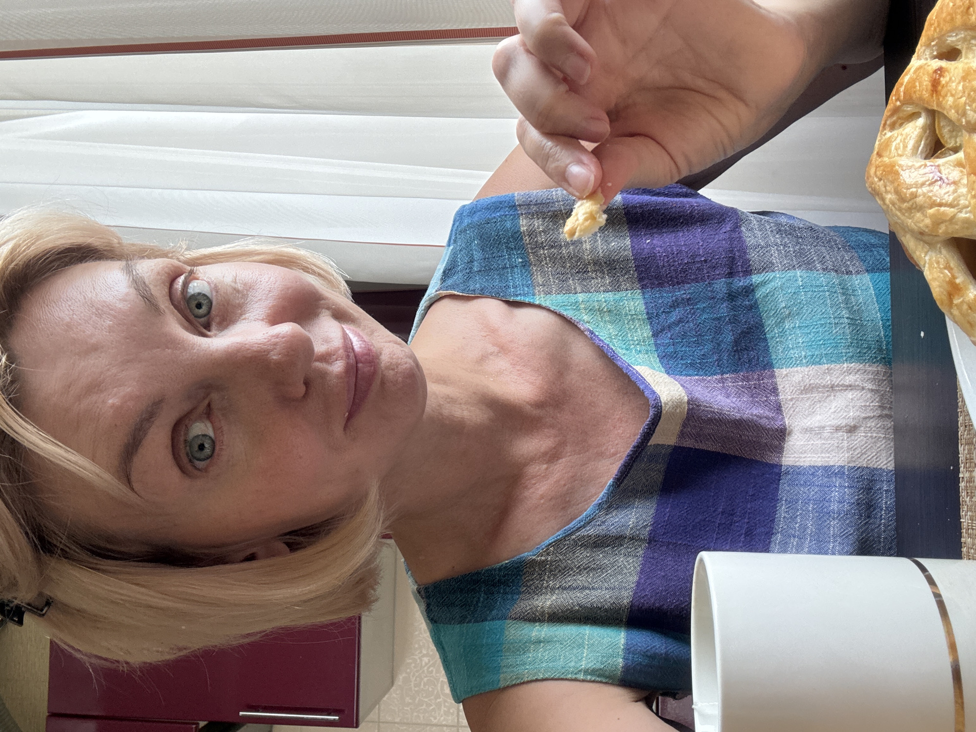 A woman with short blonde hair, wearing a blue, purple, and white checkered dress, is holding a piece of food in her right hand and looking at the camera. She has light skin and green eyes. A white mug with a gold stripe is on a dark surface to her left, and a pastry is on a white plate in the foreground. Behind her, a window with white curtains is visible, and a portion of a dark red cabinet is on the left. The setting appears to be a home environment in Sochi, Russia.
