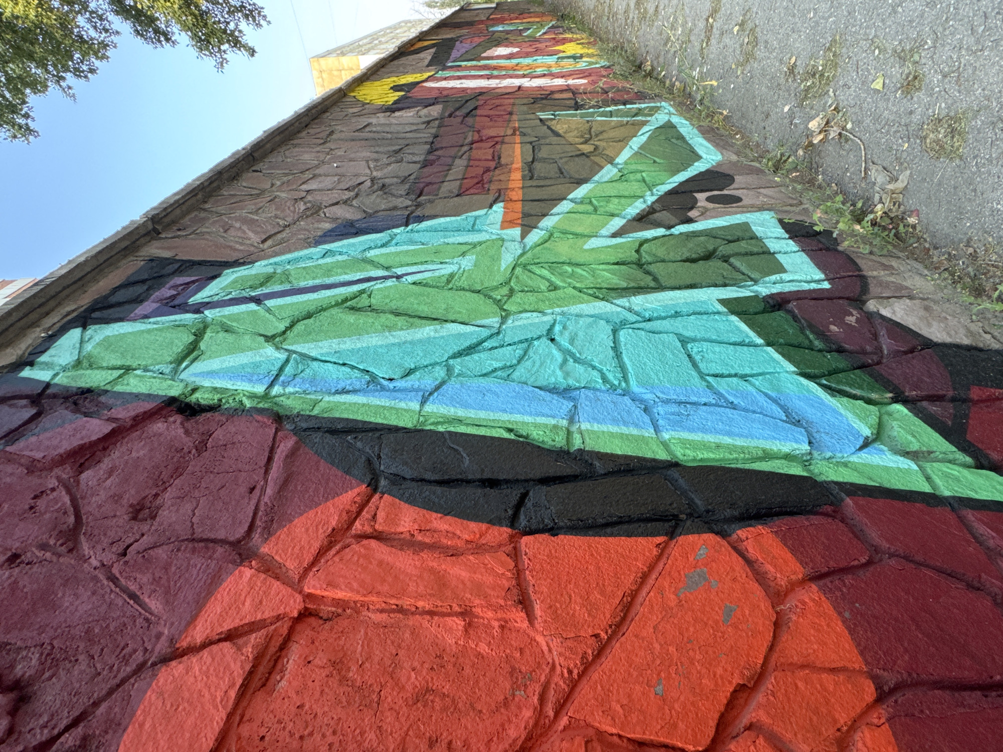 The image shows a section of a graffiti-covered stone wall in Ufa, Russia. The wall is an outdoor structure, likely bordering a street or pathway. The graffiti features bold, angular designs in vibrant colors, including shades of red, orange, black, green, and blue. The artwork is layered and appears to be in progress or partially faded in some areas. The stone texture of the wall is visible beneath the paint, with individual stones of varying sizes and irregular shapes. A paved pathway runs along the base of the wall, with some dry leaves and sparse vegetation growing between the wall and the path. In the background, a clear blue sky is visible, with the upper branches of a green tree extending into the frame from the right. A portion of a building with a lighter colored facade is also visible in the distance.