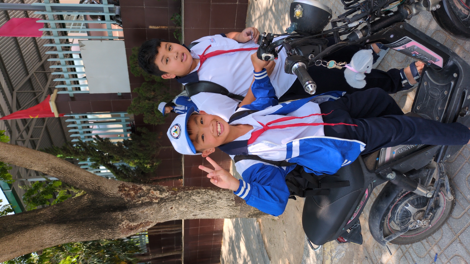 Two boys in white and blue school uniforms are on a black scooter in Lạc Viên A, Vietnam. The boy in front wears a white and blue hat and makes a peace sign. Both boys wear red scarves. The scooter has an “Osaka” sticker. A tree is visible behind them, and a Vietnamese flag is in the background. The building behind the tree has brown brick.