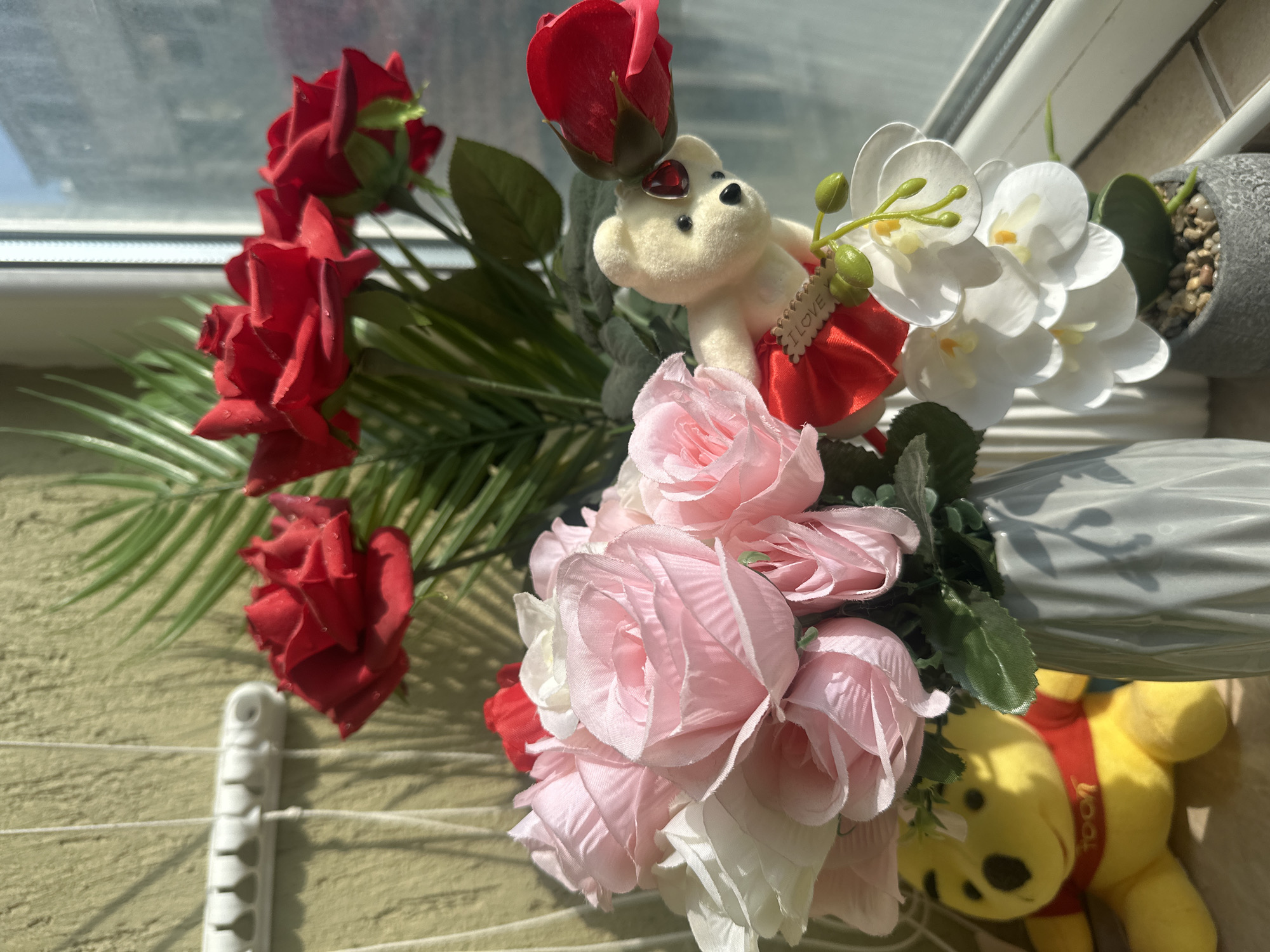 A vase of artificial flowers, including red roses, pink roses, and white orchids, is positioned near a window. A small stuffed bear wearing a red skirt and holding an "I LOVE" sign is placed among the flowers. Partially visible in the background is a yellow Winnie the Pooh plush toy and a window showing an outdoor view. The setting appears to be indoors, possibly in Varna, Bulgaria.