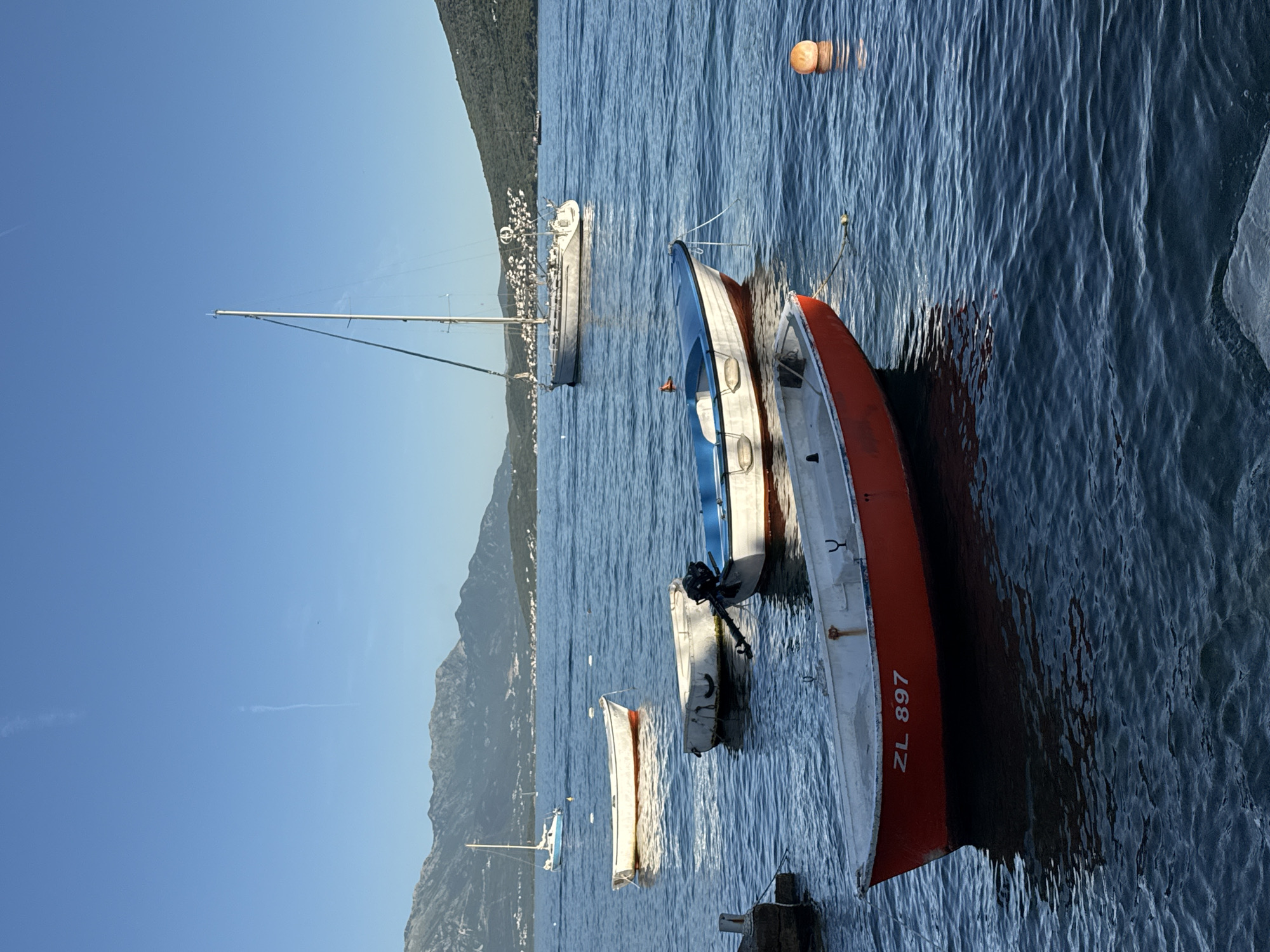 The image depicts a waterfront scene in Đenovići, Montenegro, featuring several boats moored in the water. A sailboat with a tall mast is visible in the distance. Closer to the foreground, there are several smaller boats, one painted with a blue and white hull and another with a red hull bearing the inscription "ZL 897". The water is rippled, and an orange buoy floats on the surface. Mountains are visible along the shoreline in the background under a clear blue sky. Buildings are visible on the mountainside.