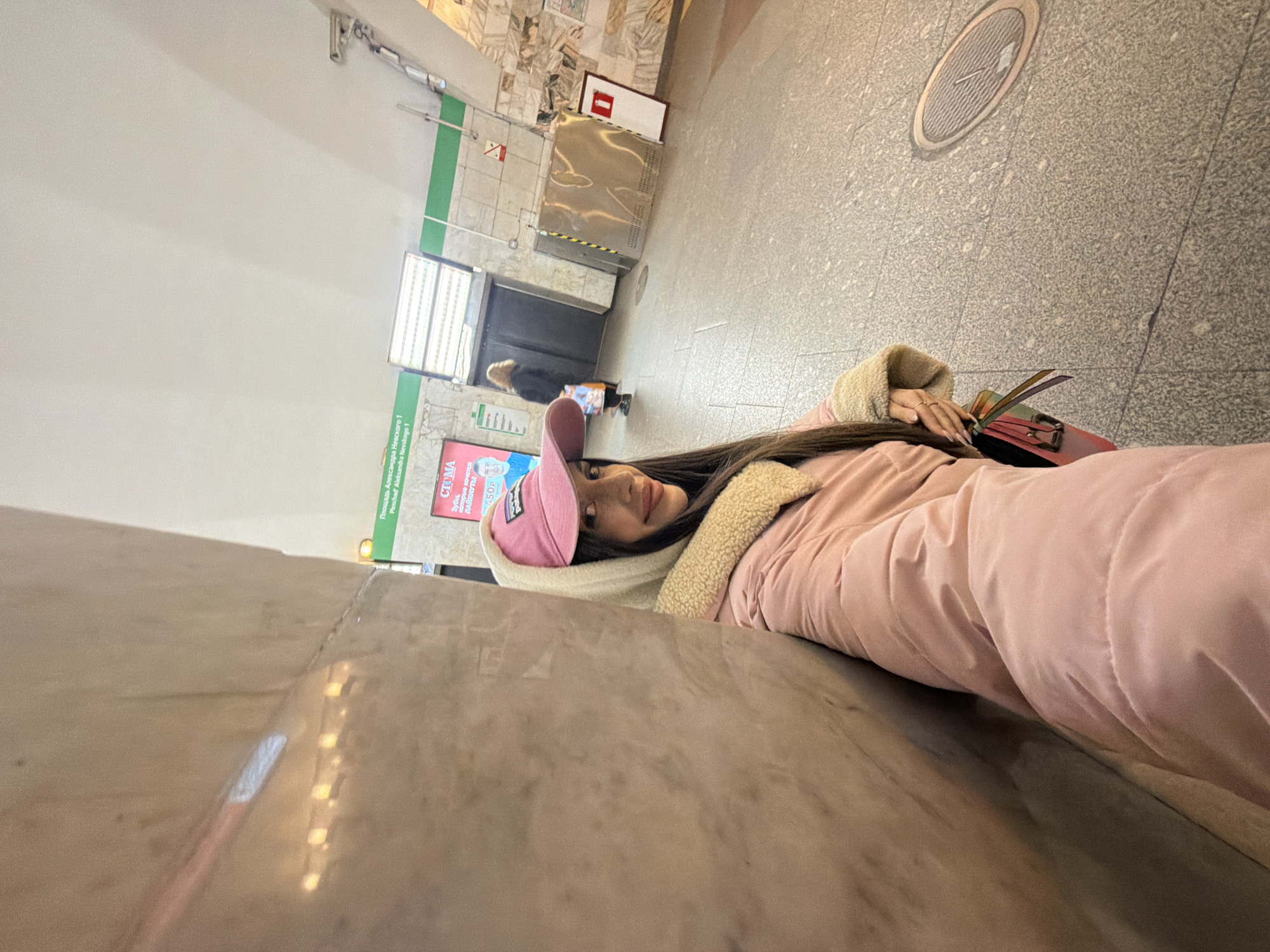A young woman is lying down on a bench in what appears to be a metro station in Saint Petersburg, Russia. She is wearing a pink jacket with a sherpa lining and a pink baseball cap. Her long brown hair is visible, and she is smiling at the camera. She has a red purse and what appears to be a wallet or book on the bench next to her.
In the background, there is an escalator, a window with advertising, and other indistinct figures in the distance. The floor is tiled. The lighting is bright, and the photo appears to have been taken with a wide-angle lens. The location is likely in a public transport station.