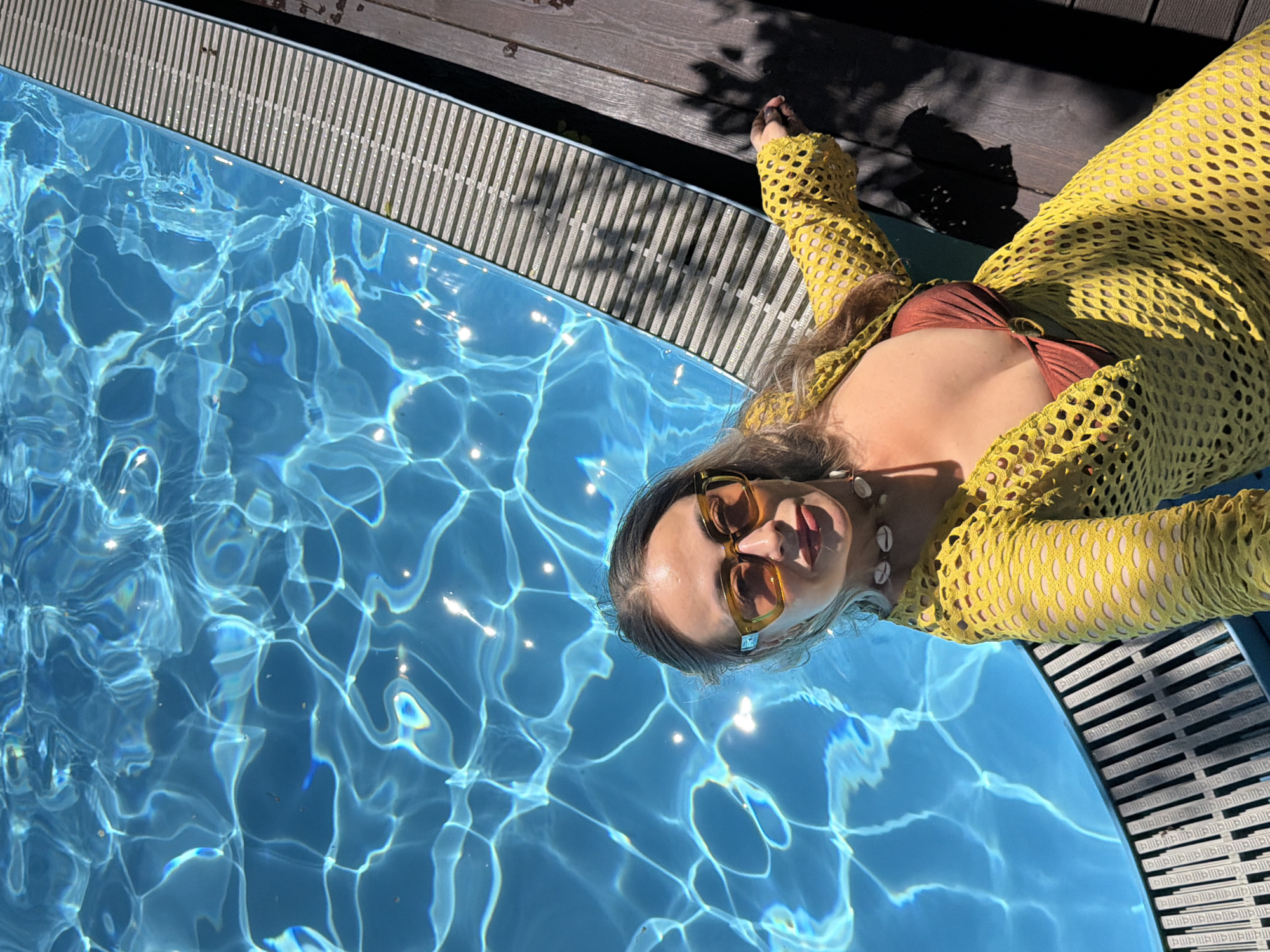 An overhead, diagonal shot captures a woman relaxing by a swimming pool in Krasnoyarsk, Russia, on a bright, sunny day. The woman, positioned at the edge of the pool, is wearing a yellow, long-sleeved mesh cover-up over a brown bikini top. She has stylish rectangular sunglasses pushed onto her nose and a seashell necklace around her neck. Her hair is light-colored and wet, suggesting she may have been in the water.
The clear blue water of the pool exhibits strong, shimmering light patterns from the sun. A patterned grate lines the edge of the pool, separating it from a dark wooden deck where parts of her arm and leg are visible, along with distinct shadows from nearby foliage or structures. The overall scene conveys a feeling of summer relaxation and leisure. No visible text is present in the image.