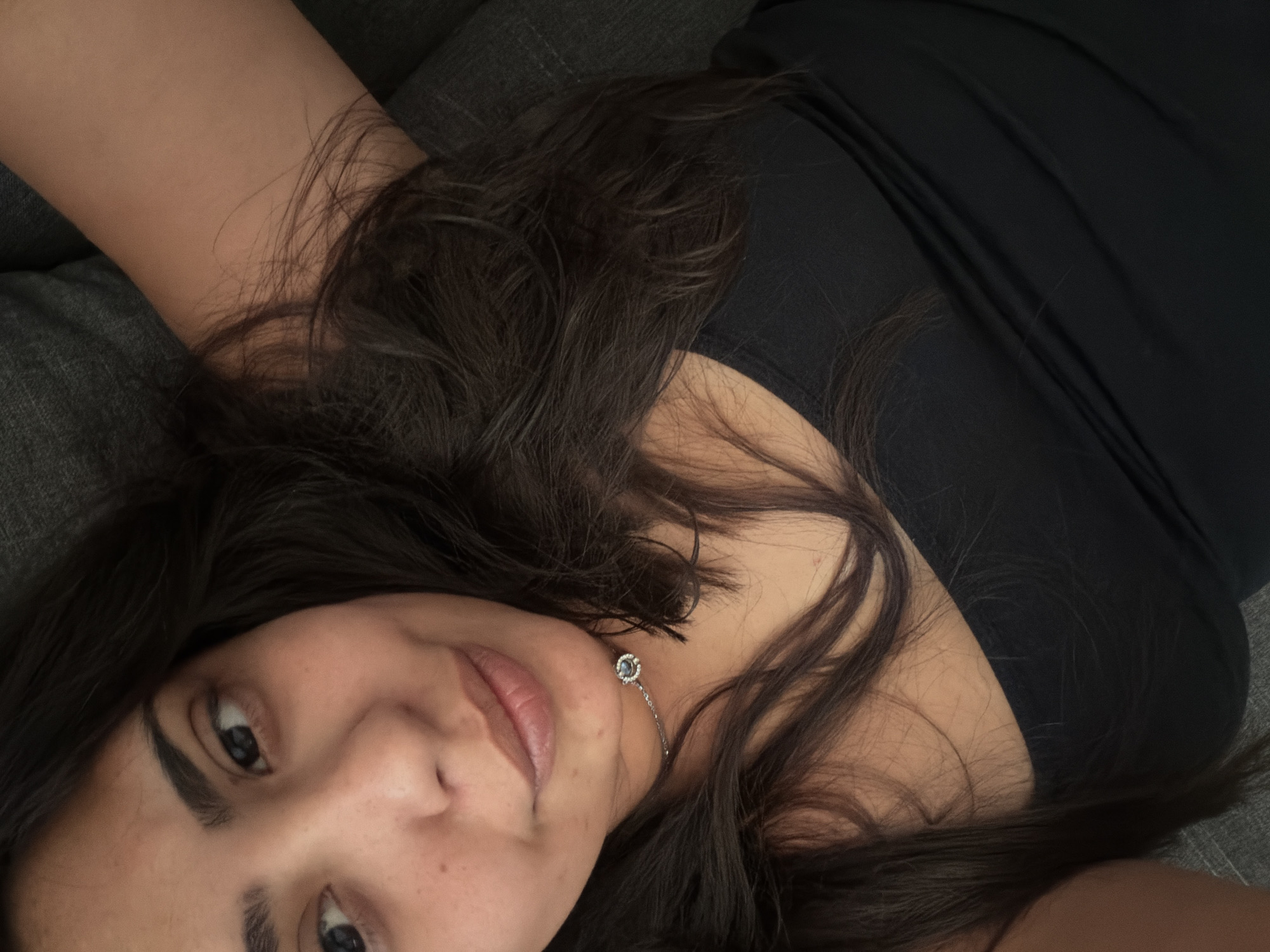 The image shows a close-up, overhead view of a person lying down, with their head tilted to the side. The person has dark hair that is spread out around their face and shoulders. They are wearing a black top. A delicate silver bracelet with a single blue gem is visible on their wrist, near their neck. The background appears to be a dark gray sofa. The image does not contain any information that would allow for the identification of a specific location in Duisburg, Germany, or any other city.