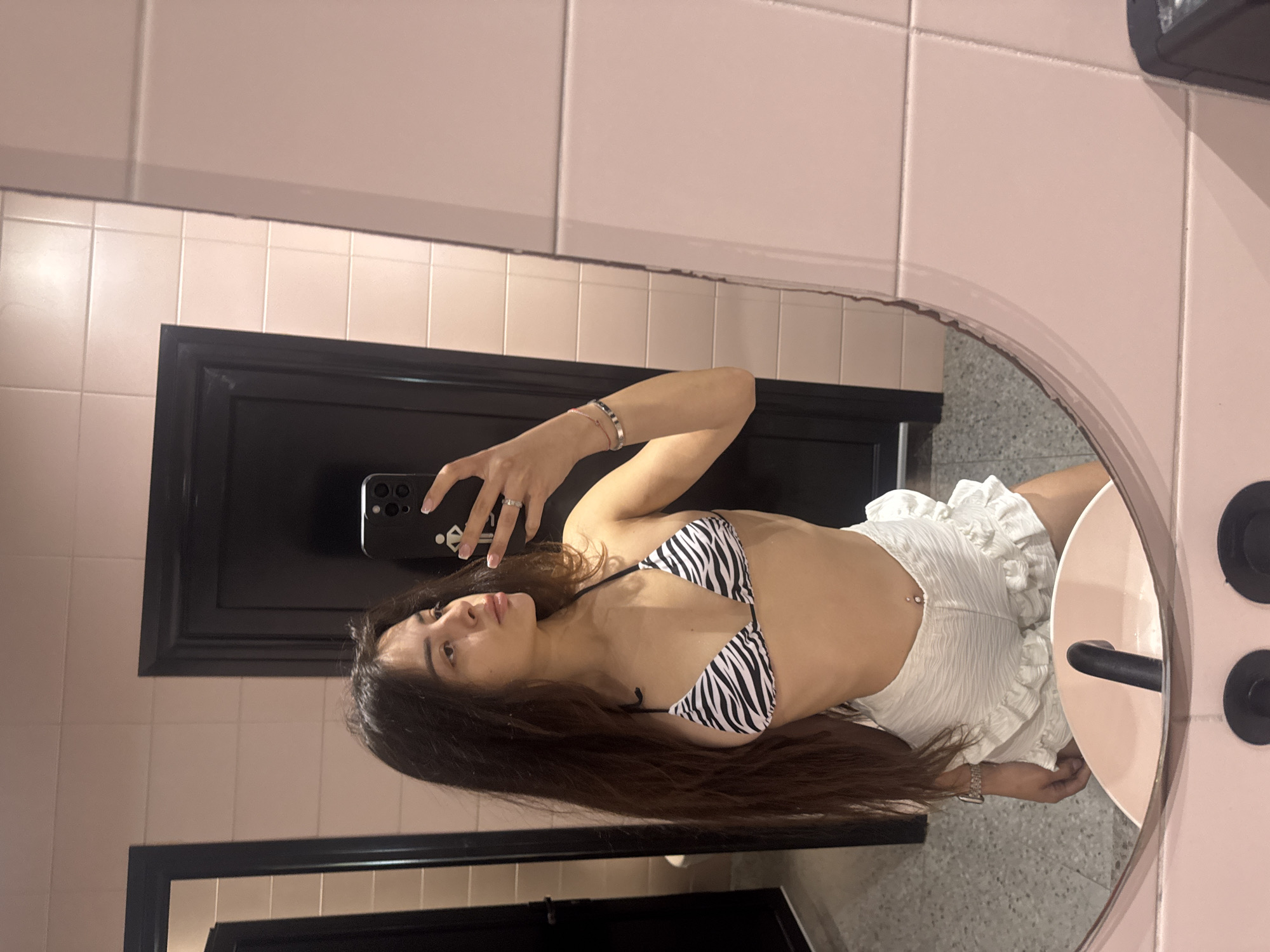 A young woman with long, dark hair is taking a mirror selfie in a restroom. She is wearing a black and white zebra-print bikini top and white ruffled shorts. Her left hand is holding a black smartphone, and her right hand is resting on her hip. A silver ring is visible on her right ring finger, and she is wearing a silver watch on her left wrist. She also has bracelets on her left wrist. A belly button piercing with a diamond stud is visible. The restroom has pale pink tiled walls and a speckled gray floor. A black door is visible in the background. The mirror is oval-shaped and framed by a pink surface. The scene is set in the city of Keramoti, Greece.