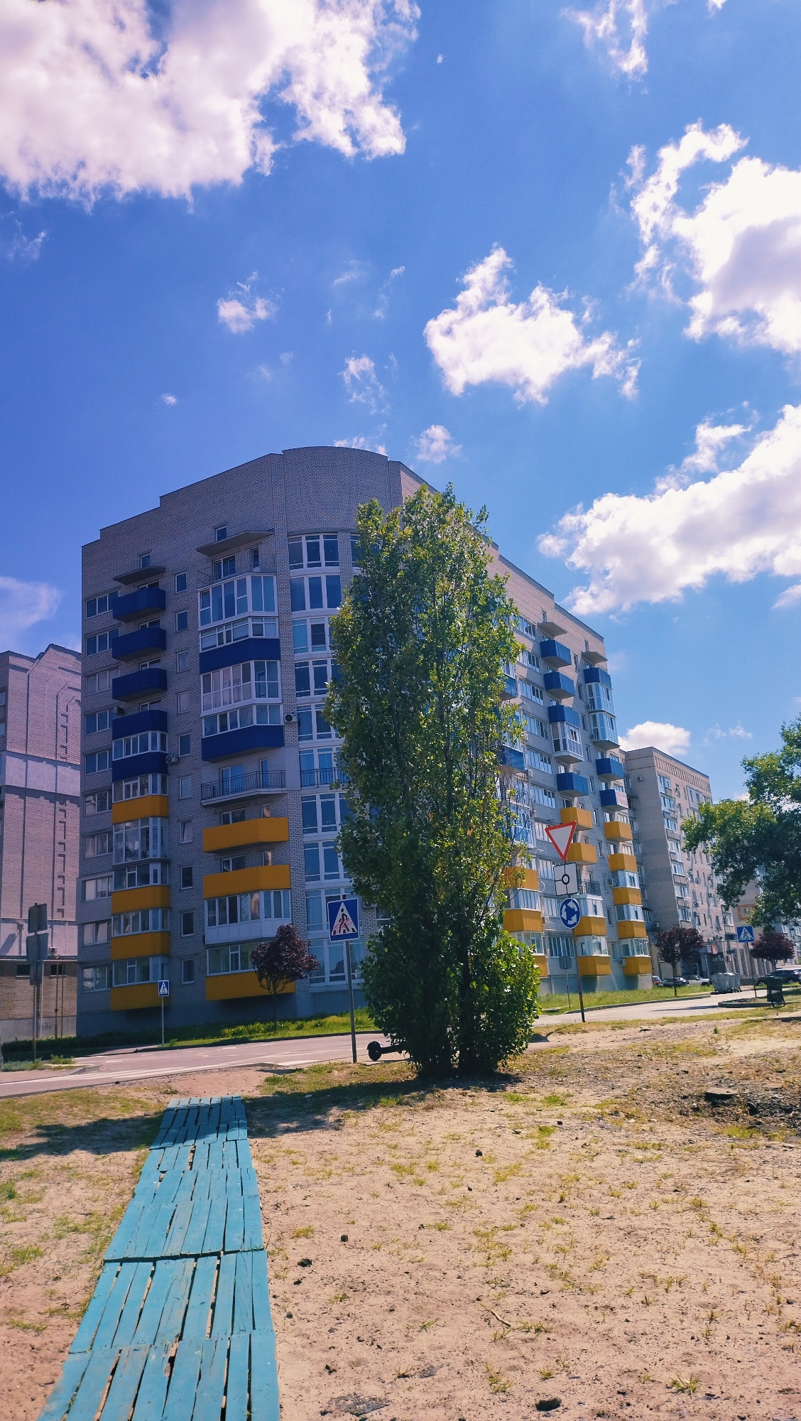 In Horishni Plavni, Ukraine, a multi-story apartment building with a curved facade dominates the scene under a bright blue sky with scattered white clouds. The building features a light grey brick exterior with alternating bands of blue and yellow balconies. A tall, slender tree stands in front of the building, partially obscuring its lower floors. The ground in the foreground is sandy with sparse green grass. A bright blue wooden walkway, made of planks, extends from the lower left corner of the frame. Road signs, including a pedestrian crossing sign and a circular traffic sign, are visible near the building. The image captures a residential urban environment on a sunny day.