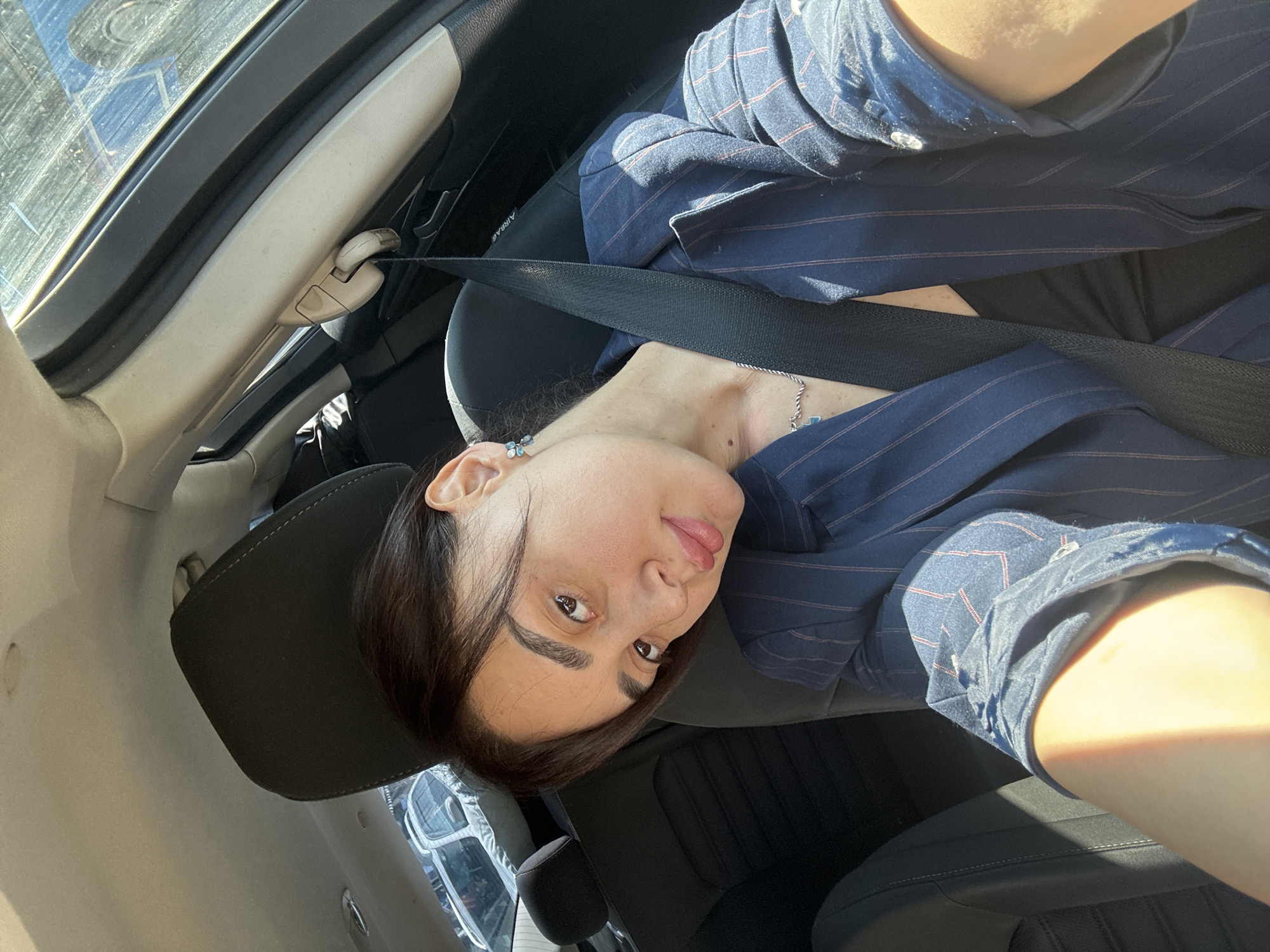 A woman is taking a selfie in a car. She is wearing a navy blue pinstripe blazer and a seatbelt. She has dark hair and is wearing earrings and a necklace. The car is parked, and a parking lot with other cars can be seen through the window. It is difficult to precisely determine the location in Tbilisi, Georgia, from this image.