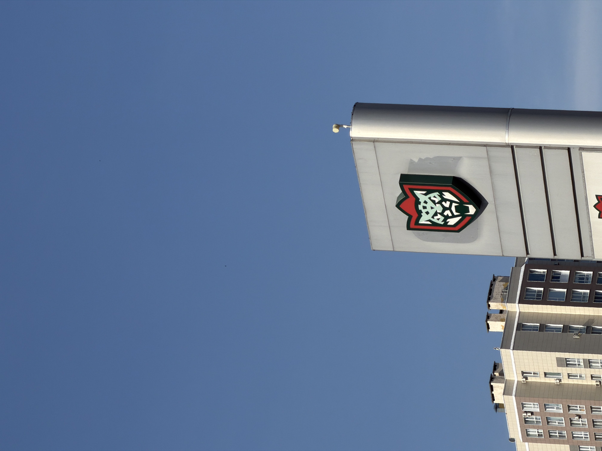 The image shows a building with a logo of Ak Bars, a professional ice hockey team based in Kazan, Russia. The logo is a stylized snow leopard head inside a shield shape with red and green colors. The building is light-colored and appears to be an office or commercial structure. A security camera is seen on the edge of the roof. A blue sky is visible behind the building. Part of an apartment building is visible near the bottom of the image.