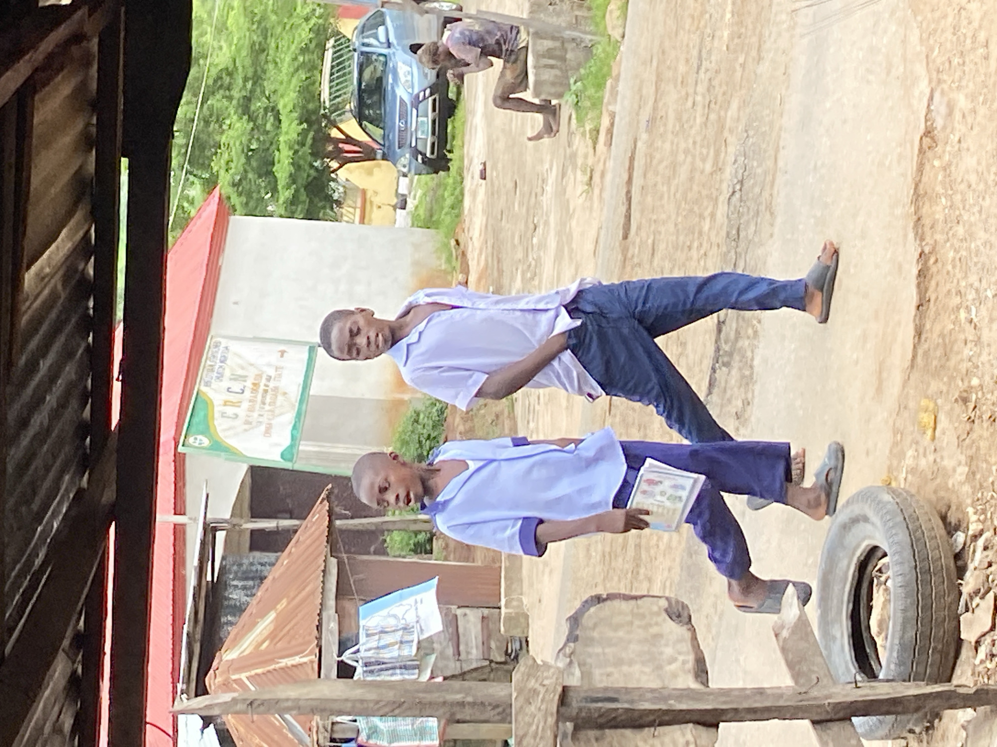 The image, taken in Mararaba, Nigeria, shows two young boys in a street scene. The boy in the foreground, wearing a light blue school uniform with purple trim, is holding a booklet and appears to be looking down at it. He has a shaved head and a curious expression. Behind him, another boy, also in a light blue school uniform, is standing with his arms outstretched, looking forward. He is wearing dark blue trousers and sandals.
The setting is outdoors, with a corrugated iron roof structure on the left of the frame. In the background, a blue vehicle is visible, along with some buildings and lush green trees. The ground is a dusty, earthy path. A large tire lies on its side in the foreground. A green sign with text is mounted on a wall in the background, partially obscured by a building. The lighting suggests it is daytime, possibly overcast given the diffused light. The overall impression is of a typical street scene in a Nigerian town, with children in school attire.