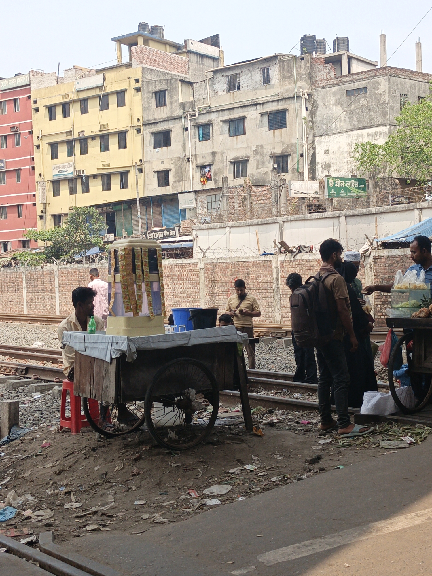 The image shows a street scene in Dhaka, Bangladesh, likely near a railway line. Several buildings of varying heights and states of repair are visible in the background. A brick wall with barbed wire tops runs parallel to the railway tracks. A food cart is positioned on the tracks, with a man seated on a red plastic stool behind it. The cart displays snacks and drinks. Several people are standing near the cart, appearing to be customers. The ground is littered with trash. A second food cart is to the right of the first, with a man serving customers. The railway tracks run through the scene.
