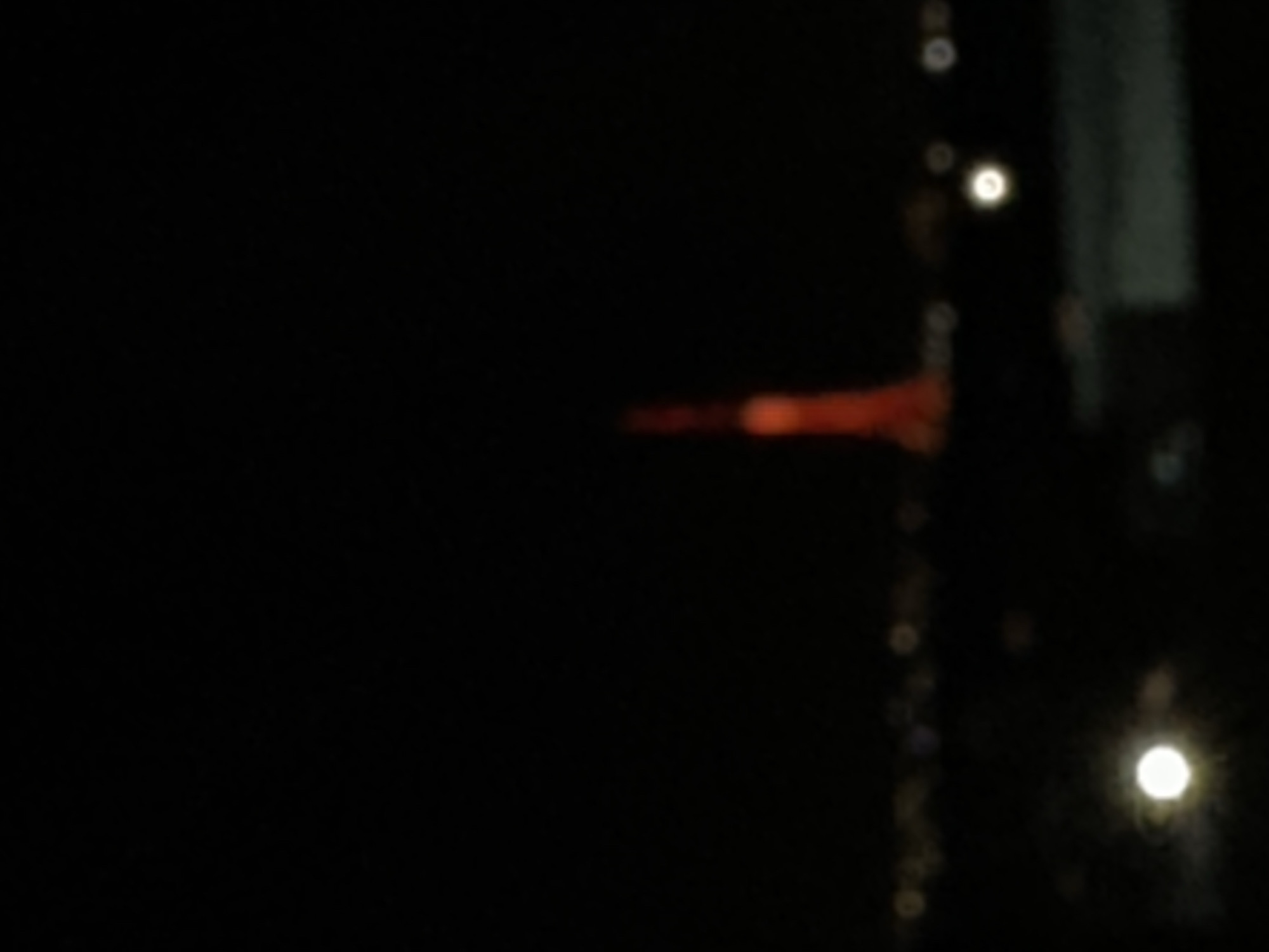 The image appears to be a nighttime shot of the Yerevan TV Tower in Yerevan, Armenia. The tower is illuminated in red light and can be seen on the right side of the image. There are also some blurry street lights in the foreground, with a dark background.
