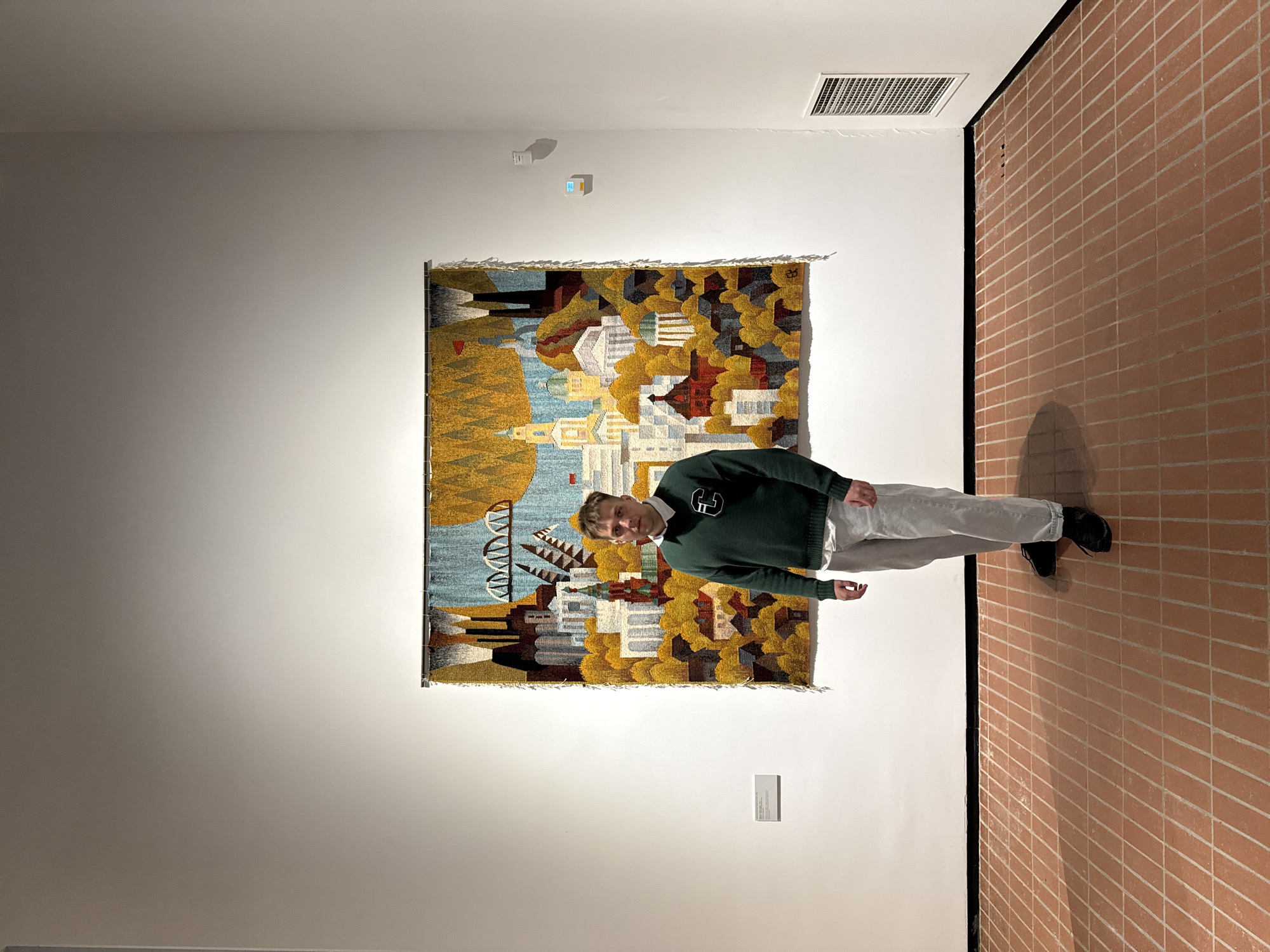 A young, blond man stands next to a large woven wall hanging. He wears a dark green sweater with a white letter "G", light gray pants, and black shoes. The tapestry depicts a city scene in shades of brown, yellow, red, and blue with stylized buildings, trees, and a bridge. Above the tapestry are two small sensors or devices affixed to the wall. To the right of the scene is a brick wall. A vent is installed in the ceiling above the brick wall. This photo was likely taken in Perm, Russia.
