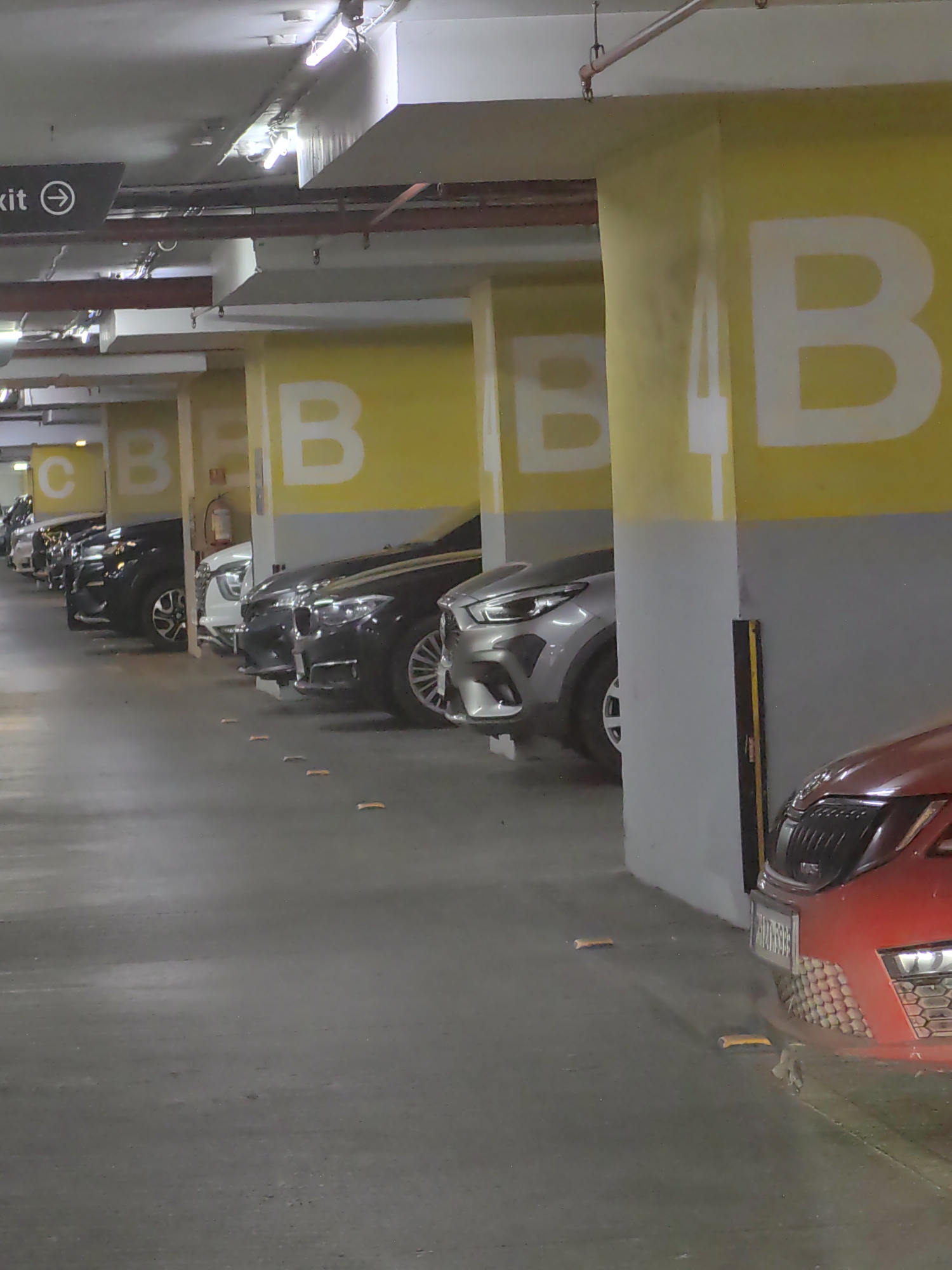 A low-angle, eye-level perspective captured inside a multi-level parking garage in Mumbai, India. The image is dominated by the concrete floor and large concrete pillars painted with yellow and grey sections. Prominently displayed on the yellow sections of the pillars are large, white letters that appear to be part of a parking zone designation, with "B" being the most visible letter.
A row of parked cars is visible, receding into the background. These vehicles are of various colors including black, white, silver, and a partial view of a red car in the foreground on the right. The cars exhibit modern designs, suggesting they are relatively new models. The overall lighting in the garage appears to be artificial, with fluorescent lights visible on the ceiling, casting a somewhat dim and even illumination across the scene.
The floor of the parking garage is a smooth concrete surface, showing some scuff marks and a few small, reflective markers on the ground that likely aid in guiding vehicles. The pillars have a textured finish, and one of them has a dark, vertical protective strip along its edge. In the upper left of the image, a black sign with an arrow pointing to the right is partially visible, with the word "exit" appearing to be on it, indicating an egress point. The environment is clean and orderly, typical of a well-maintained parking facility. There are no people visible in the frame.