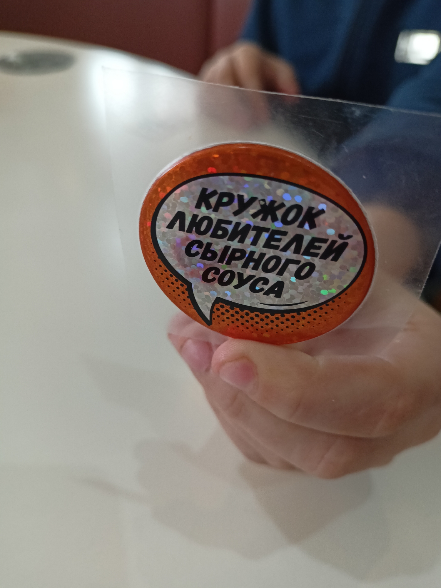 The image shows a close-up of a hand holding a transparent plastic sheet. Adhered to the plastic sheet is a small, round sticker with an orange border and a halftone pattern. The sticker features a stylized speech bubble shape containing text in Russian: "КРУЖОК ЛЮБИТЕЛЕЙ СЫРНОГО СОУСА", which translates to "Club of Cheese Sauce Lovers." The sticker has a holographic pattern inside the text area. The background shows a white table and a person wearing a blue garment. The scene is located in Novosibirsk, Russia.