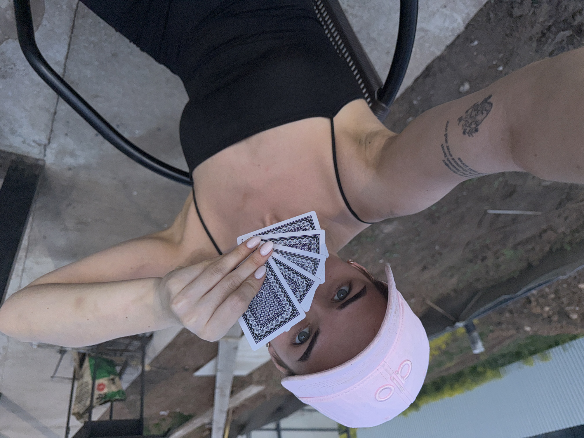 A woman wearing a black top and a pink cap is holding playing cards in front of her face. She has a tattoo on her left arm. She is sitting in a black metal chair outdoors. The location is Лобаново, Russia. The background includes gravel, vegetation, and a fence.