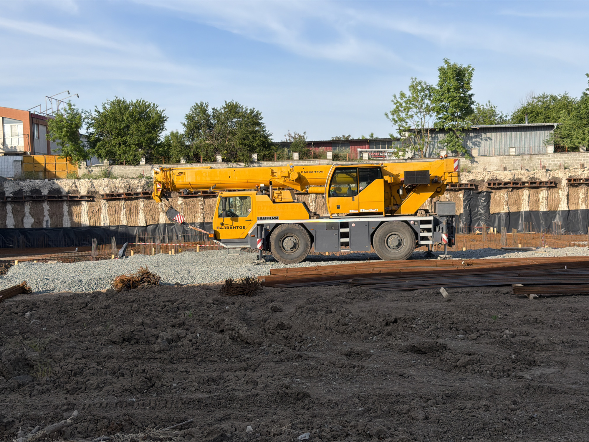 The image depicts a construction site in Burgas, Bulgaria. A yellow Liebherr mobile crane is positioned on the site. In the foreground, there is disturbed soil, piles of rebar, and gravel. In the background, a partially excavated area is visible, reinforced with steel beams and covered with black plastic sheeting. Buildings and trees are visible in the background. The sky is partly cloudy. There are no people visible in the image.