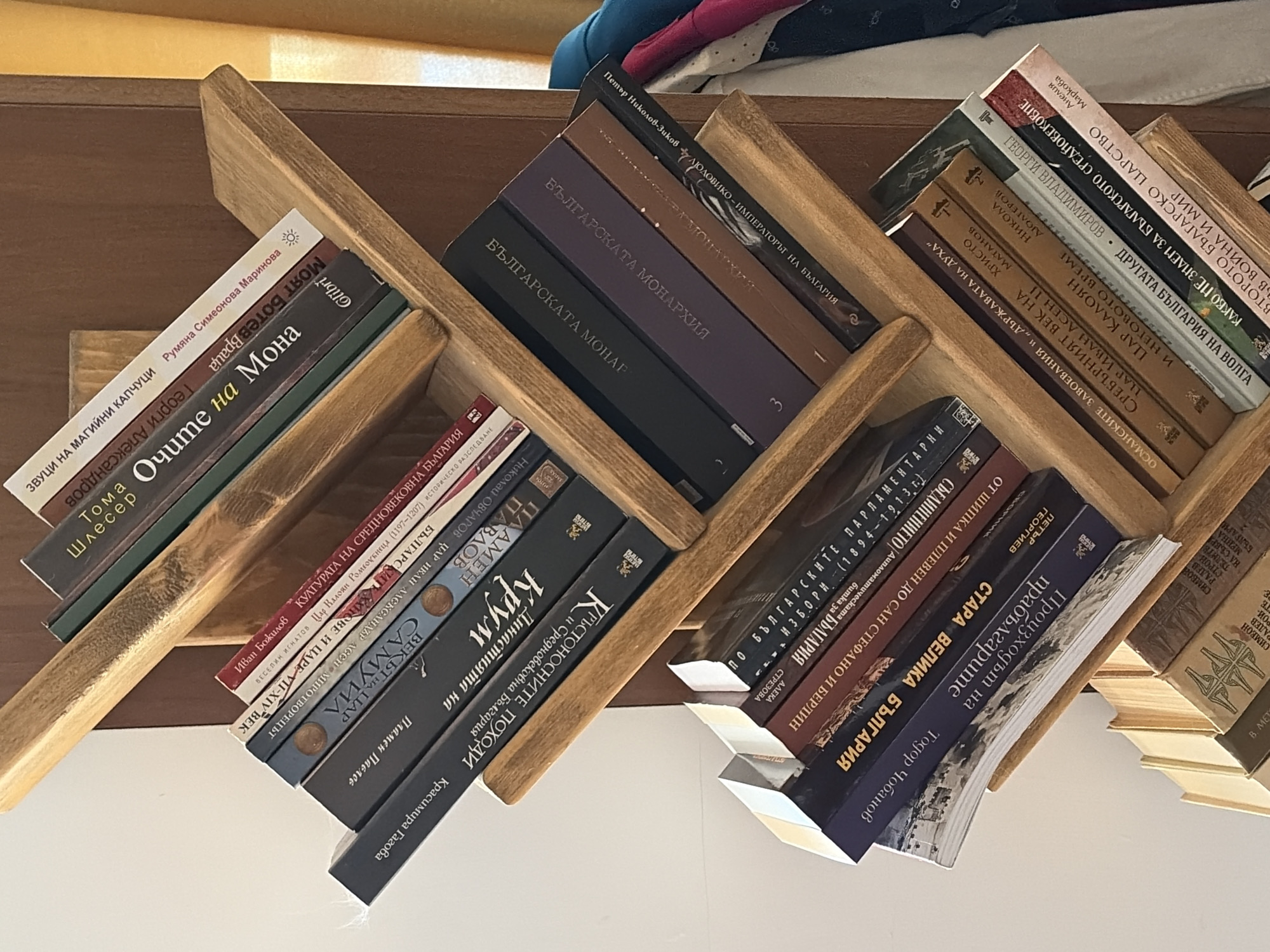 The image shows a wooden, tiered bookshelf filled with books. The bookshelf is leaning against a wall, with a yellow curtain visible on the right. The books are of various sizes and colors, with some titles and authors visible. The overall impression is of a home library or a small bookstore. There are no people or animals present in the image. The lighting suggests it is daytime. The specific location in Vratsa, Bulgaria cannot be determined from the image alone.