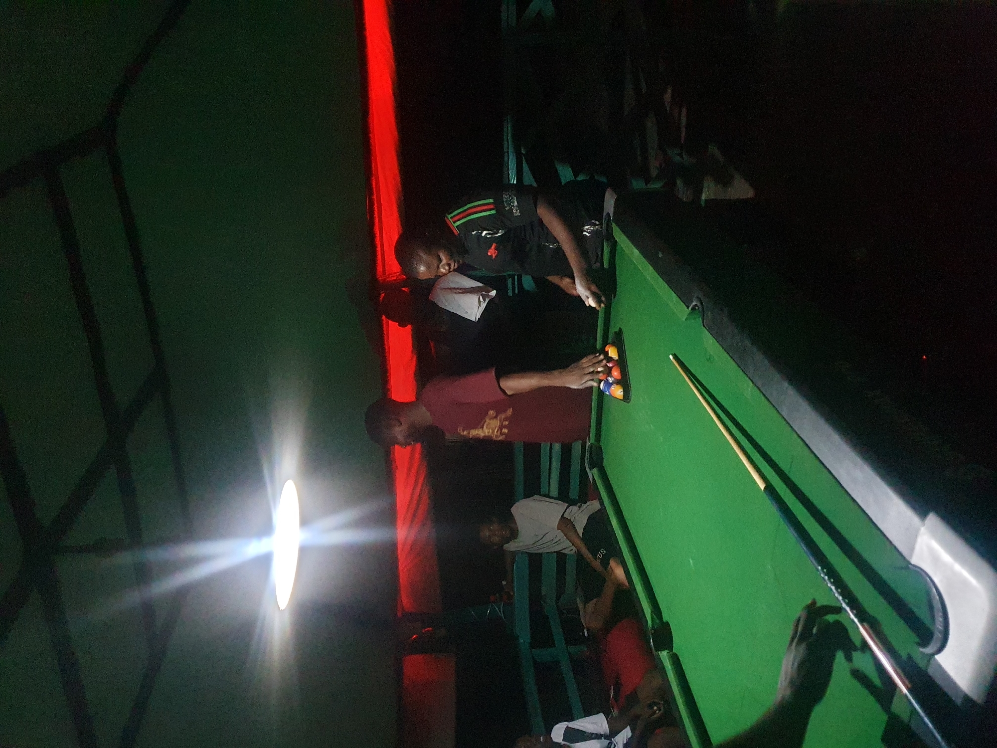 In Lokoja, Nigeria, a group of individuals gathers around a green billiards table under the illumination of a bright overhead light. The scene is set outdoors, possibly at a bar or recreational area, with a dark, possibly corrugated roof structure overhead. Red fabric or material is visible in the background, creating a contrasting ambiance.
Three men and one woman are the prominent figures in the image. One man, wearing a maroon t-shirt, stands near the table, seemingly engaged in conversation. Another man, in a grey t-shirt, is seated at the edge of the table. A third man, dressed in a black and green striped shirt, is actively holding a white billiards ball, positioned as if about to play. A fourth individual, partially visible on the left, wears a white and black outfit.
The billiards table is set with several colored balls, arranged in a rack. A cue stick lies across the table, indicating a game in progress or about to commence. The overall atmosphere appears casual and social, with the individuals interacting around the game. The lighting casts strong highlights and shadows, contributing to a somewhat dramatic visual effect. The environment suggests a relaxed evening spent playing billiards.