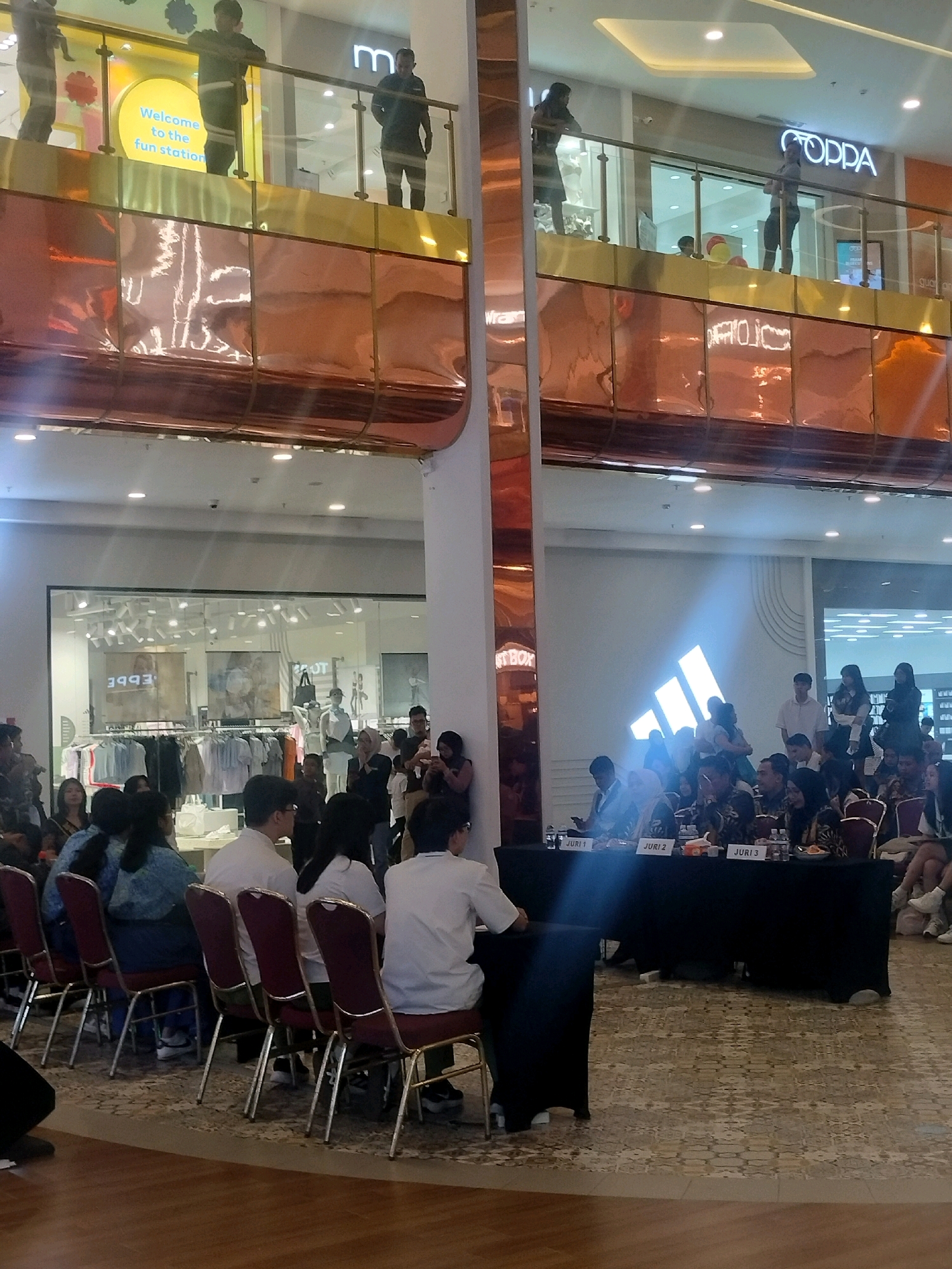 The image depicts an indoor event, likely a competition or presentation, taking place in a modern shopping mall environment in Batam, Indonesia. The foreground features a row of seated individuals, some of whom appear to be judges, positioned behind a long black table draped with a fabric cover. Labels indicating "JURI 1", "JURI 2", and "JURI 3" are visible on the table, confirming the presence of judges. The individuals in this section are dressed in various attire, with some wearing formal shirts.
Behind the judges, a group of onlookers are standing and observing the proceedings. The background reveals a brightly lit storefront, possibly a clothing store, with racks of garments displayed behind glass windows. Signs with mirrored text, one reading "EPPE" and another potentially "STBOX", are visible on the storefront.
Further up in the mall, on an upper level, several individuals are seen leaning on a railing. One person stands near a sign that reads "Welcome to the fun station". Another area on the upper level shows a store with the prominent "COPPA" logo. The architecture of the mall is modern, with glass railings and reflective surfaces, suggesting a commercial setting designed for shopping and entertainment. The lighting is bright and diffuse, typical of indoor retail spaces. The overall scene suggests a public event in progress within a commercial establishment in Batam.