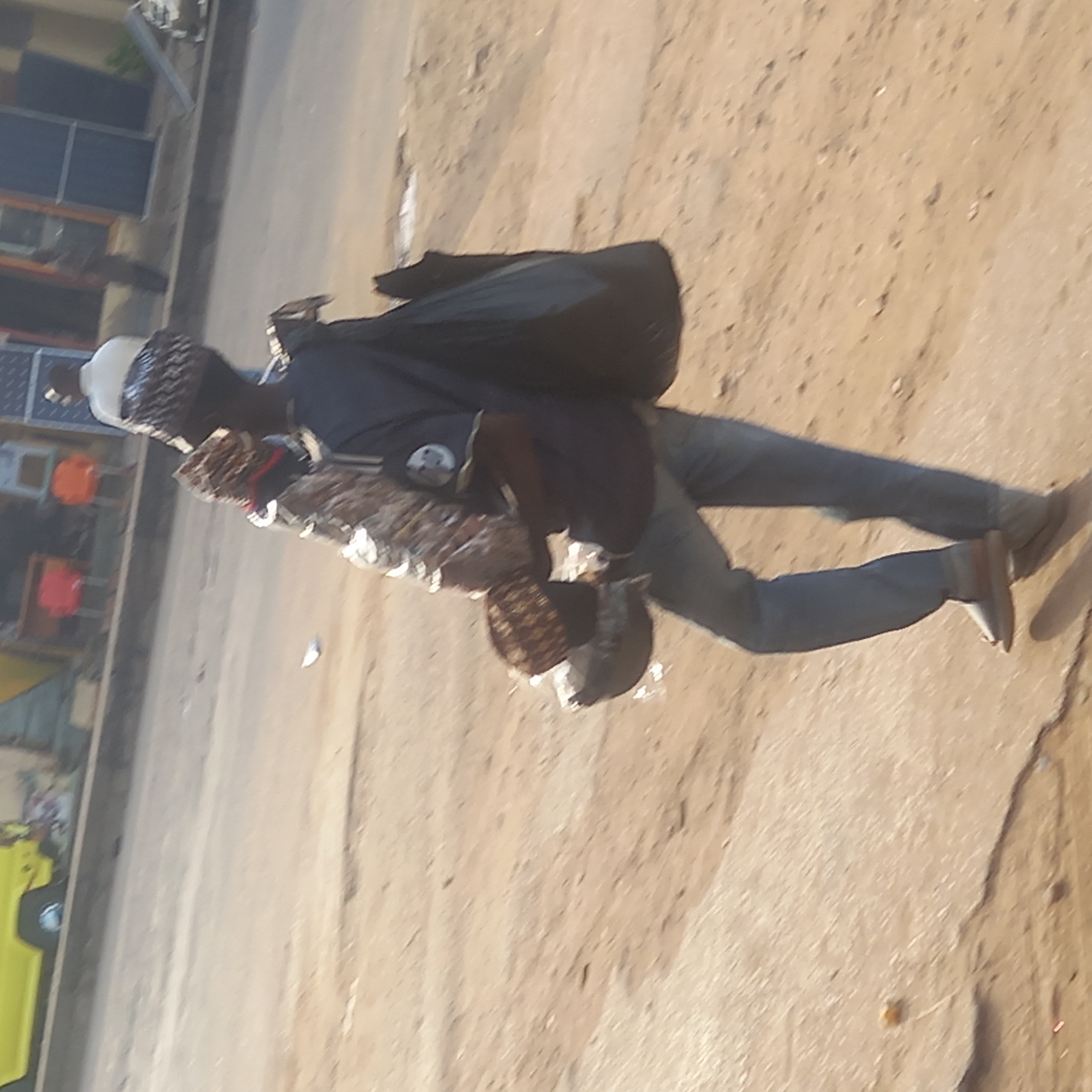 An individual in Dadi Riba, Nigeria, is seen walking on a street. The person wears a white cap, a dark blue shirt, blue jeans, and brown shoes, and carries a black bag on their back. They also carry what appears to be wicker baskets or similar items in their arms. The environment is a city street with buildings visible in the background.