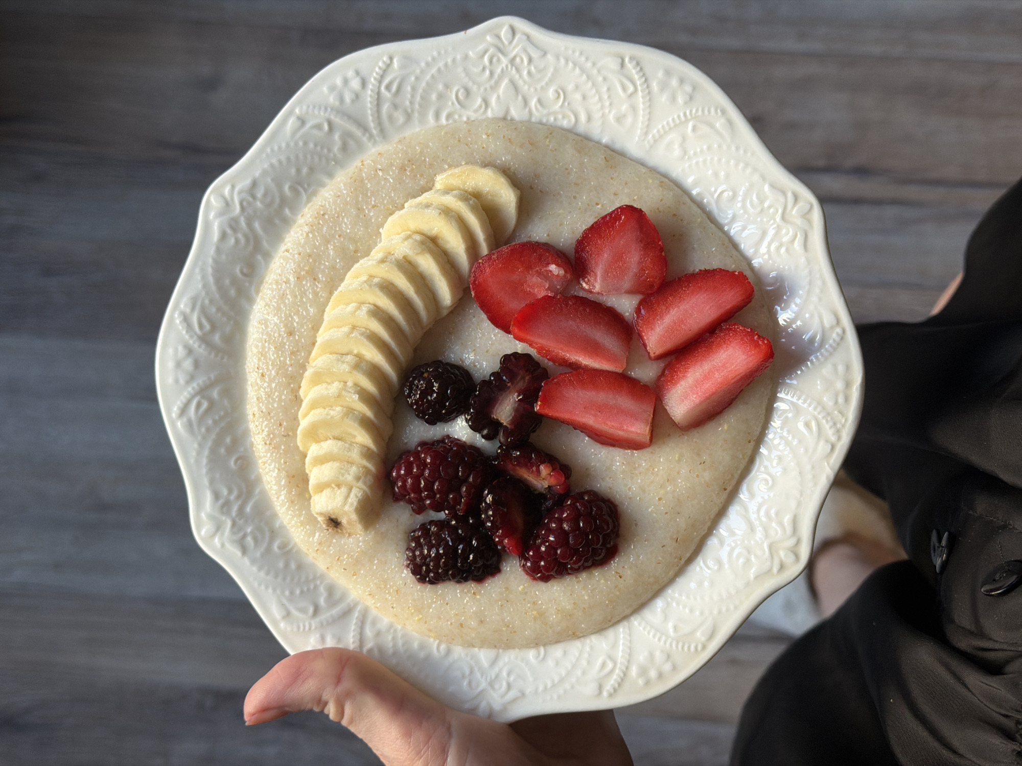 A top-down shot shows a white, ornate plate held in a hand, presumably for a person about to eat. The plate contains a smooth, beige substance, likely cream of wheat or grits. Arranged on top are a line of sliced bananas, several sliced strawberries, and a cluster of dark blackberries. The background is a wooden surface, and a small portion of a person wearing black clothing is visible on the right side of the frame. The location is not identifiable from the image.