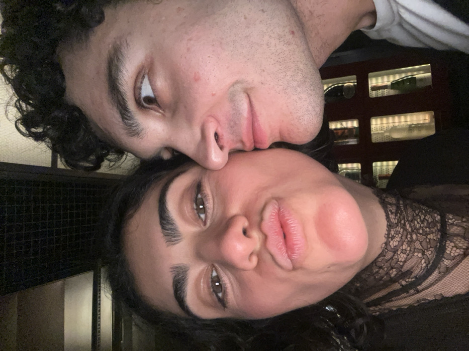 A dark, tilted close-up shot features a couple. The man, positioned above, has curly dark hair and a slightly stubbled face. His gaze is angled towards the viewer. Below him, the woman pouts her lips, and her eyes are directed at the camera. She has dark hair and prominent eyebrows. A sheer, patterned garment adorns her neckline. Behind them, a dimly lit, grid-like structure with glowing compartments containing bottles is visible.