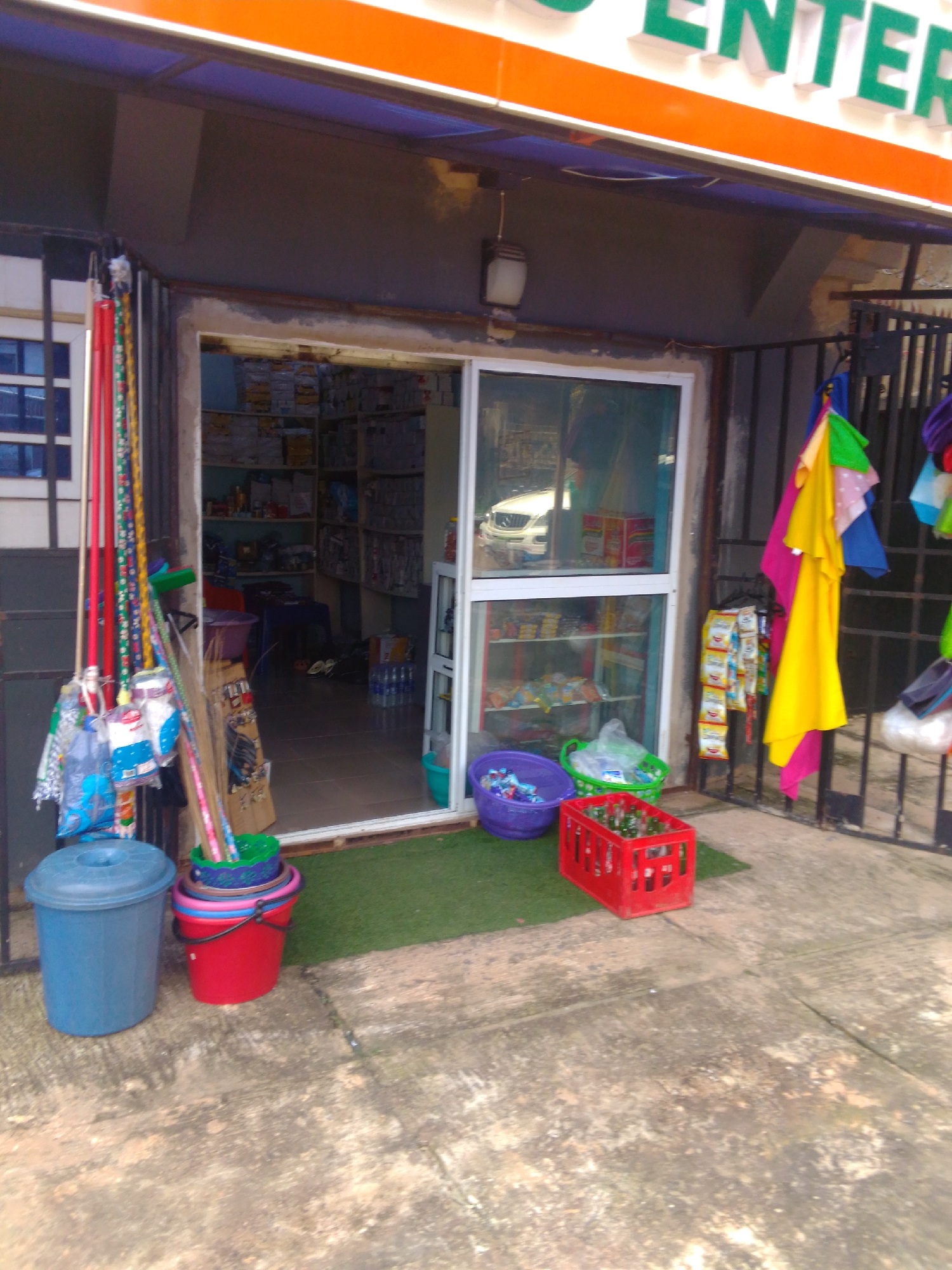 This image depicts a small storefront in Enugu, Nigeria, likely a convenience store or small supermarket, judging by the merchandise displayed inside and outside. The storefront features a wide entrance with a glass door and window, showcasing shelves stocked with various packaged goods. Outside the entrance, cleaning supplies such as brooms and buckets are arranged, along with colorful plastic containers and a red crate filled with bottles. Several racks of snacks or small food items are hung on the right side of the storefront. The building's exterior is characterized by a prominent orange and green signage that reads "ENTER". The overall scene suggests a bustling commercial area.
