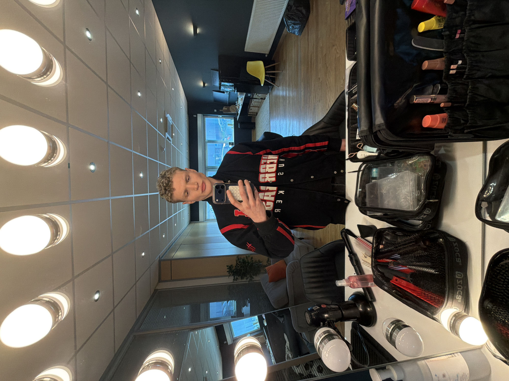 A young man is taking a selfie in front of a vanity mirror, presumably in a dressing room or backstage area, likely in Trafford, United Kingdom given the prompt. The room has a grid-patterned ceiling with circular lights. A makeup artist's kit and various hair styling tools are arranged on the vanity. Behind the man's reflection, there is a seating area with chairs and a plant visible through the reflection of a window. The man is wearing a black jacket with red stripes.