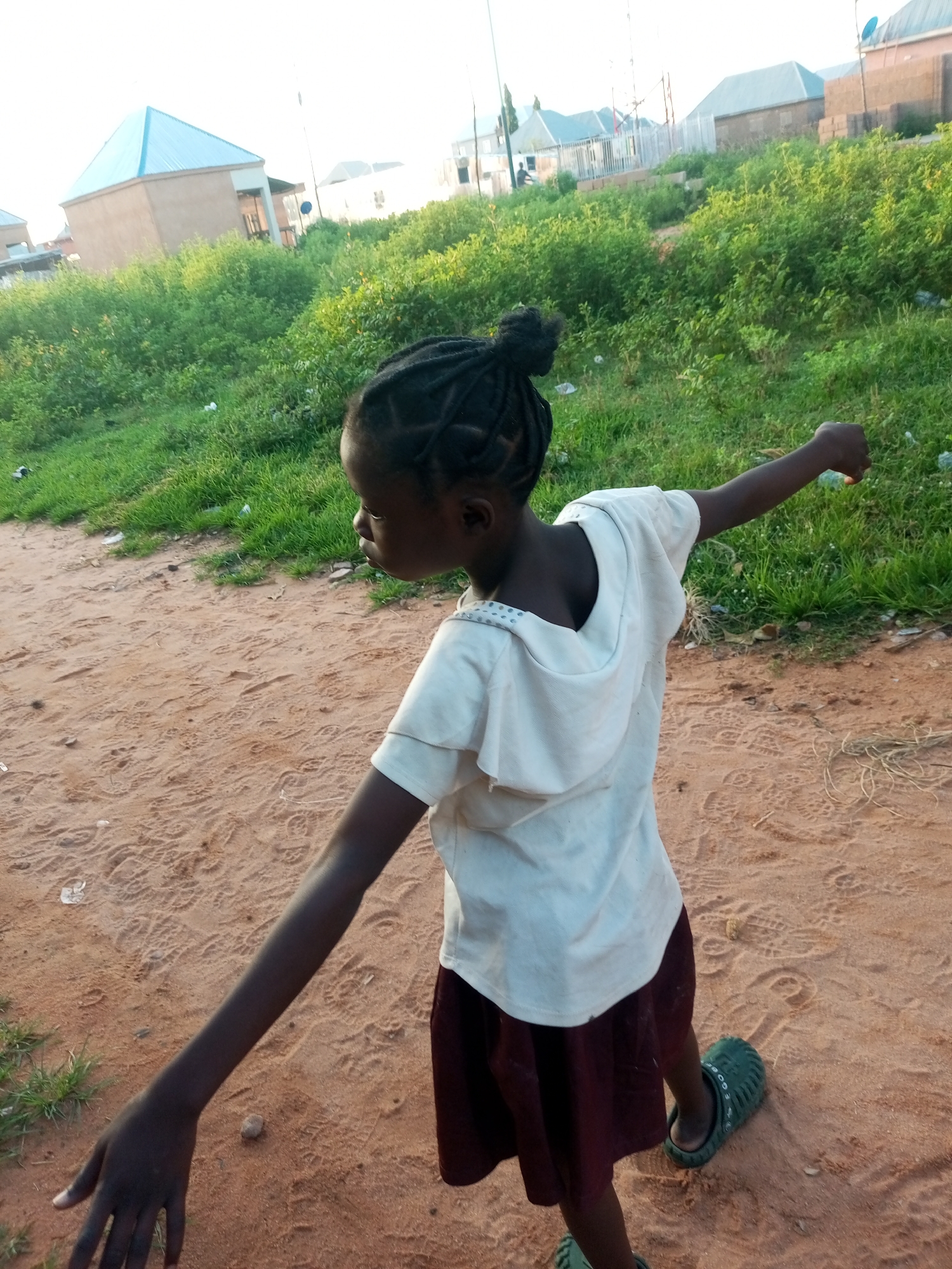 A young girl with dark skin and braided hair stands on a sandy path in Tempure, Nigeria. She is wearing a light-colored, short-sleeved shirt and a dark skirt. Her left arm is extended to her side, and her right arm is bent at the elbow, with her fist clenched. She is looking away from the camera. The background shows a grassy area with some buildings and trees. The sky is bright, suggesting it is daytime.