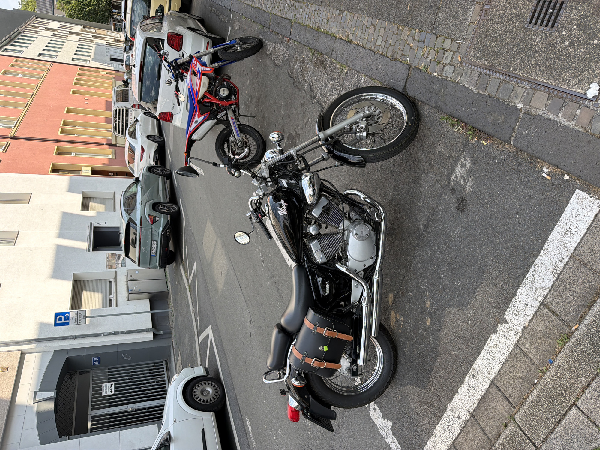 A black Yamaha motorcycle with leather saddlebags is parked on a street in Bochum, Germany, next to a white Volkswagen car and a red and white dirt bike. The street is lined with buildings, and there are other cars and motorcycles parked in the background. A blue parking sign with German text is visible on the sidewalk. The scene appears to be in a residential or commercial area with paved sidewalks and asphalt road. There are no people visible in the image. The lighting suggests it is daytime, possibly late afternoon due to the shadows.