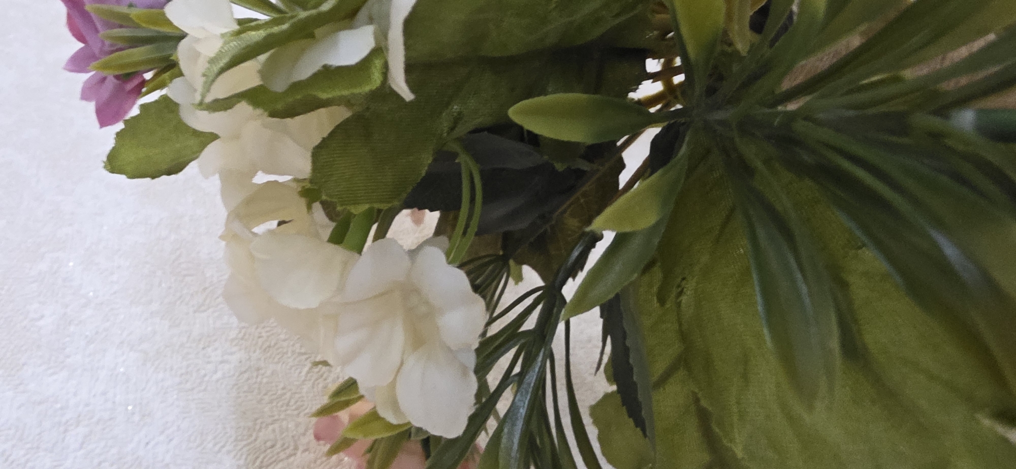 The image shows a close-up view of a floral arrangement. The arrangement features several white flowers with ruffled petals, as well as some pink and purple flowers. The flowers are interspersed with green leaves and stems. The background is a textured, light-colored surface, possibly a wall. There is no indication of the city of Sumgayit, Azerbaijan, or any people in the image.