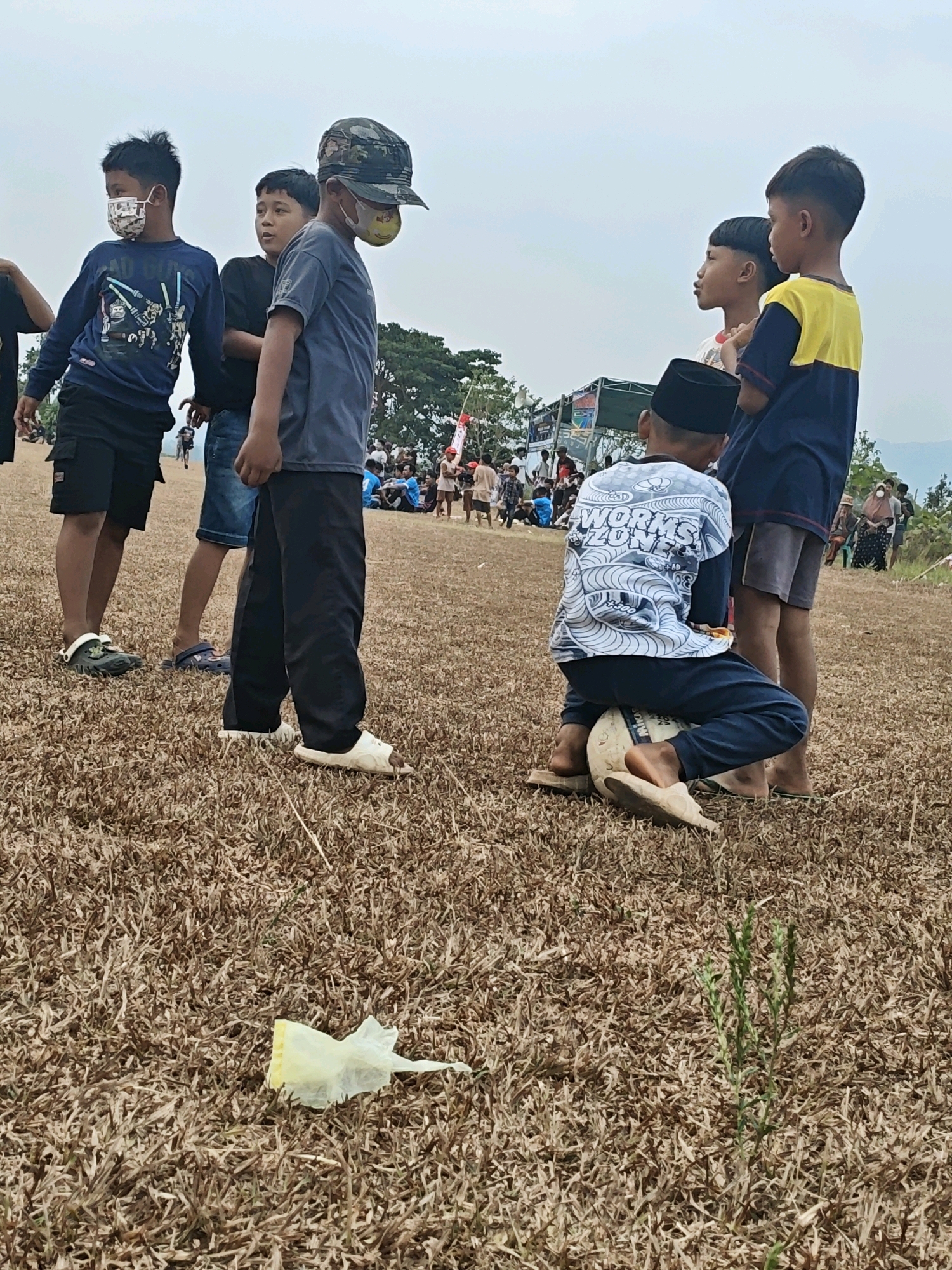 This image depicts a group of boys on a dry, grassy field in Karang Sari, Indonesia. The primary focus is on five young boys, some wearing face masks. In the foreground, a boy wearing a black peci hat and a white long-sleeved shirt with a "Worms Zone" graphic is sitting on a soccer ball. To his right, another boy in a yellow and dark blue t-shirt stands. To the left of the boy on the ball, three other boys are gathered. One, wearing a camouflage cap and a light blue t-shirt, stands looking forward. Another boy to his left, in a dark blue long-sleeved shirt with a graphic of characters, is also facing forward. A third boy, in the background and partially obscured, is wearing a black t-shirt and denim shorts. In the background, a crowd of people are visible, as well as tents and structures, suggesting an event or gathering. The environment is characterized by dry, brown grass covering the ground, with some sparse greenery. The sky is overcast. A crumpled yellow piece of plastic is visible on the ground in the lower left corner.