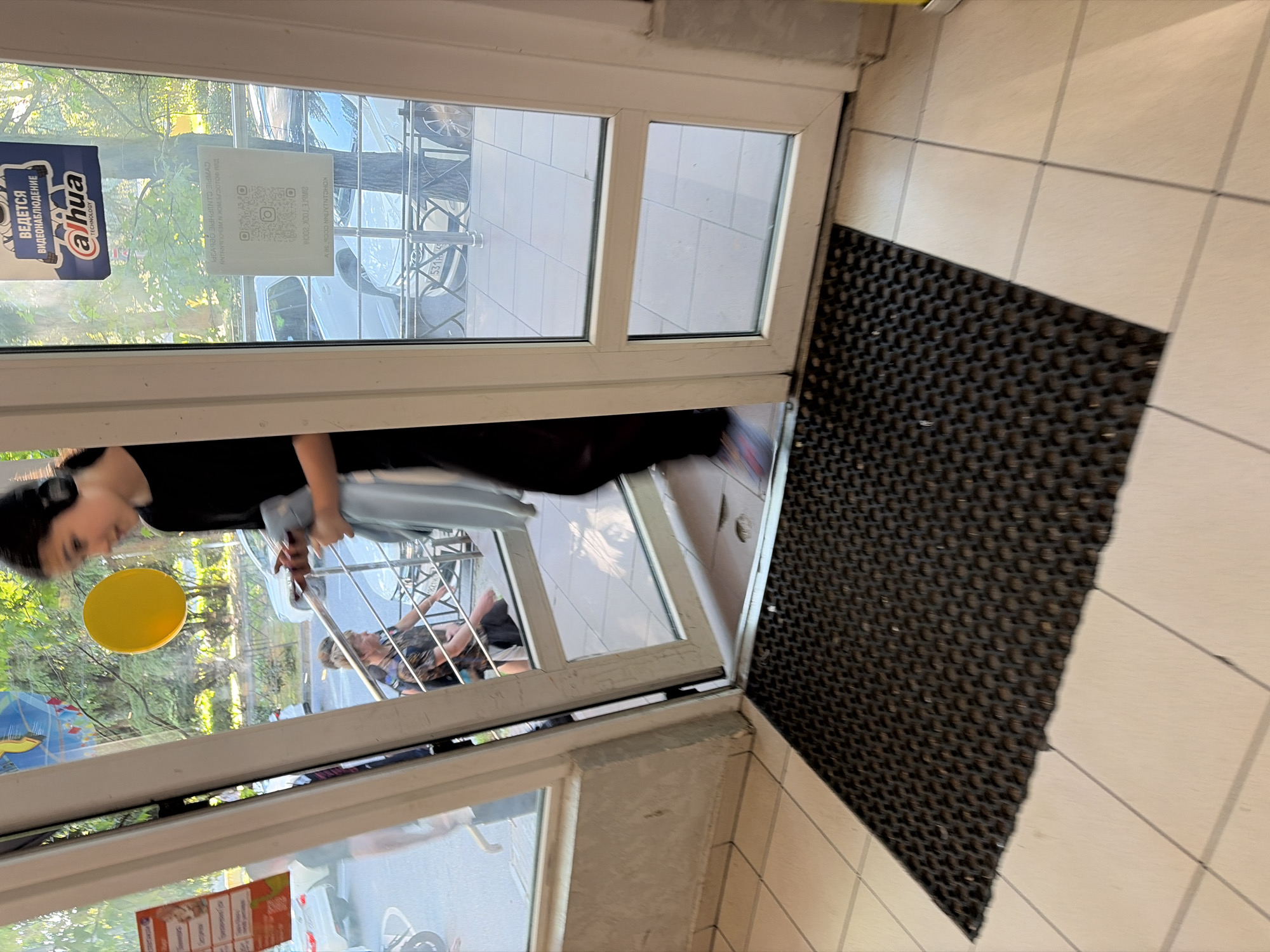 The image depicts an interior view looking out through a white-framed glass door onto an urban outdoor scene in Sochi, Russia. A young individual, likely female, wearing black attire and black headphones, is in motion, stepping through the partially open left door panel. She holds a smartphone in her right hand and a light blue garment in her left hand, with a multi-colored sneaker visible on her right foot.
Inside, the floor is tiled with light-colored square ceramic tiles and a black, honeycomb-patterned rubber mat is positioned directly in front of the doorway. The wall to the left of the door is a plain light grey.
On the closed right glass panel of the door, two signs are affixed. The upper sign is a blue rectangle with white Cyrillic text indicating "Video surveillance is being conducted," accompanied by the "Dahua Technology" logo. Below it, a white rectangular sticker displays a QR code and additional Cyrillic text, including "СОБЛЮДАЙТЕ ДИСТАНЦИЮ И НОСИТЕ МАСКУ" (Maintain distance and wear a mask), with some text appearing mirrored due to reflections.
Beyond the door, an outdoor environment is visible under bright daylight. A second person, an adult with light hair and wearing a patterned shirt, is seated or leaning near a metal railing that separates a paved area from a street or parking zone. Several white cars are parked, one displaying a partially visible Russian license plate "531 H 95." Green trees and foliage are present in the background, and a distinct yellow circular object is visible outside through the glass. A faint reflection of another person is visible on the inner surface of the left door panel.