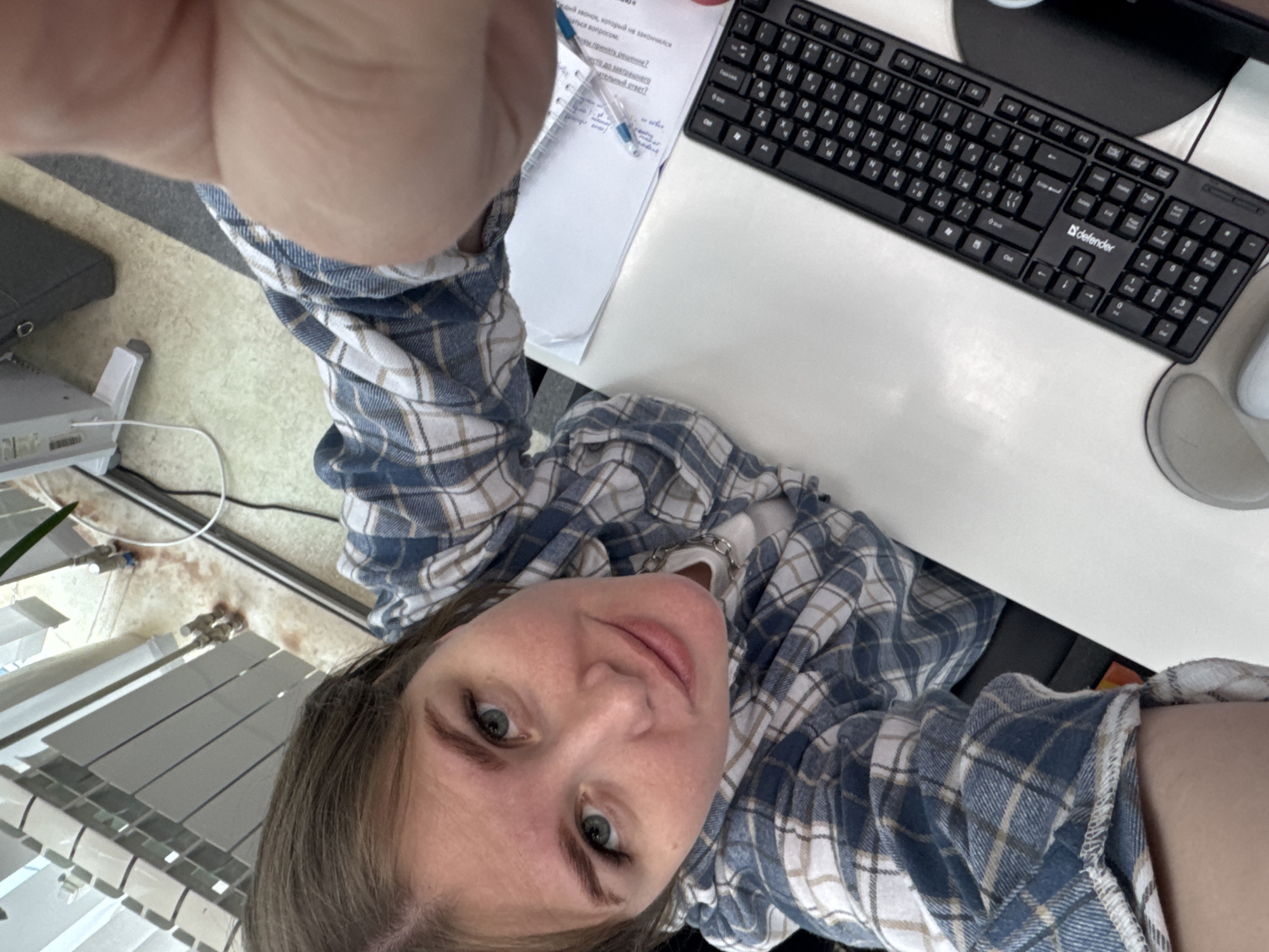 A woman in a blue and white plaid shirt poses for a selfie at a desk. The desk contains a Defender brand keyboard, a computer monitor, and a piece of paper with text in Russian, including questions and the words "Оковы принять решение?" and "тельный ответ?". She is holding a pen. A window with blinds is visible to the left. The location appears to be in Perm, Russia, based on the Russian text.