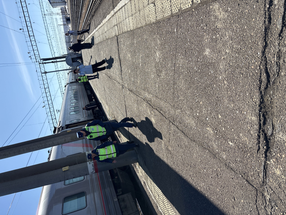 The image depicts a train station platform in Kropachyovo, Russia, under a clear blue sky. A silver passenger train, displaying a red Russian Railways logo, is stationary alongside the platform on the left. Two individuals in dark blue uniforms with bright neon-green reflective vests and dark caps are walking towards the foreground on the asphalt platform. Long shadows extend from them and from two prominent dark grey utility poles located between them and the train. One utility pole features a yellow triangular warning sign. Further down the platform, several other individuals are visible; one wears a light blue jacket and another a neon-green vest. The platform surface exhibits cracks and minor deterioration, with yellow tactile paving strips visible along its edge. In the background, multiple sets of train tracks are visible, crossed by an extensive overhead catenary system supported by metal gantries. Distant structures are faintly visible on the horizon to the right.