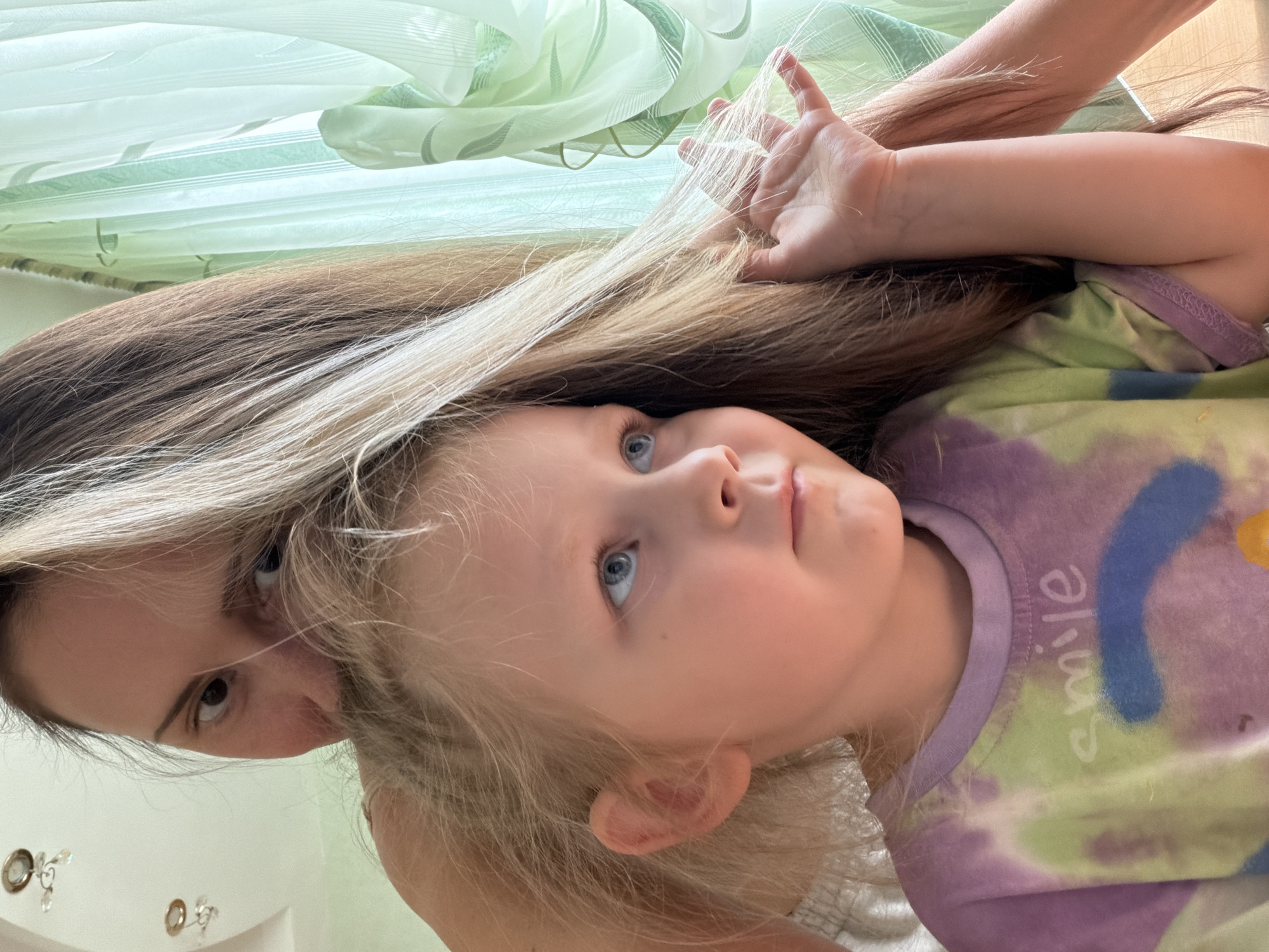 A selfie shows a fair-skinned woman with light brown hair looking down at the camera from the left side of the frame. Partially obscured by the woman's hair is a fair-skinned child with blonde hair and blue eyes, looking upward. The child wears a shirt with a purple neckline and tie-dye pattern with the word "smile" written on it. The background consists of a light green sheer curtain. The location is Kamyshin, Russia.