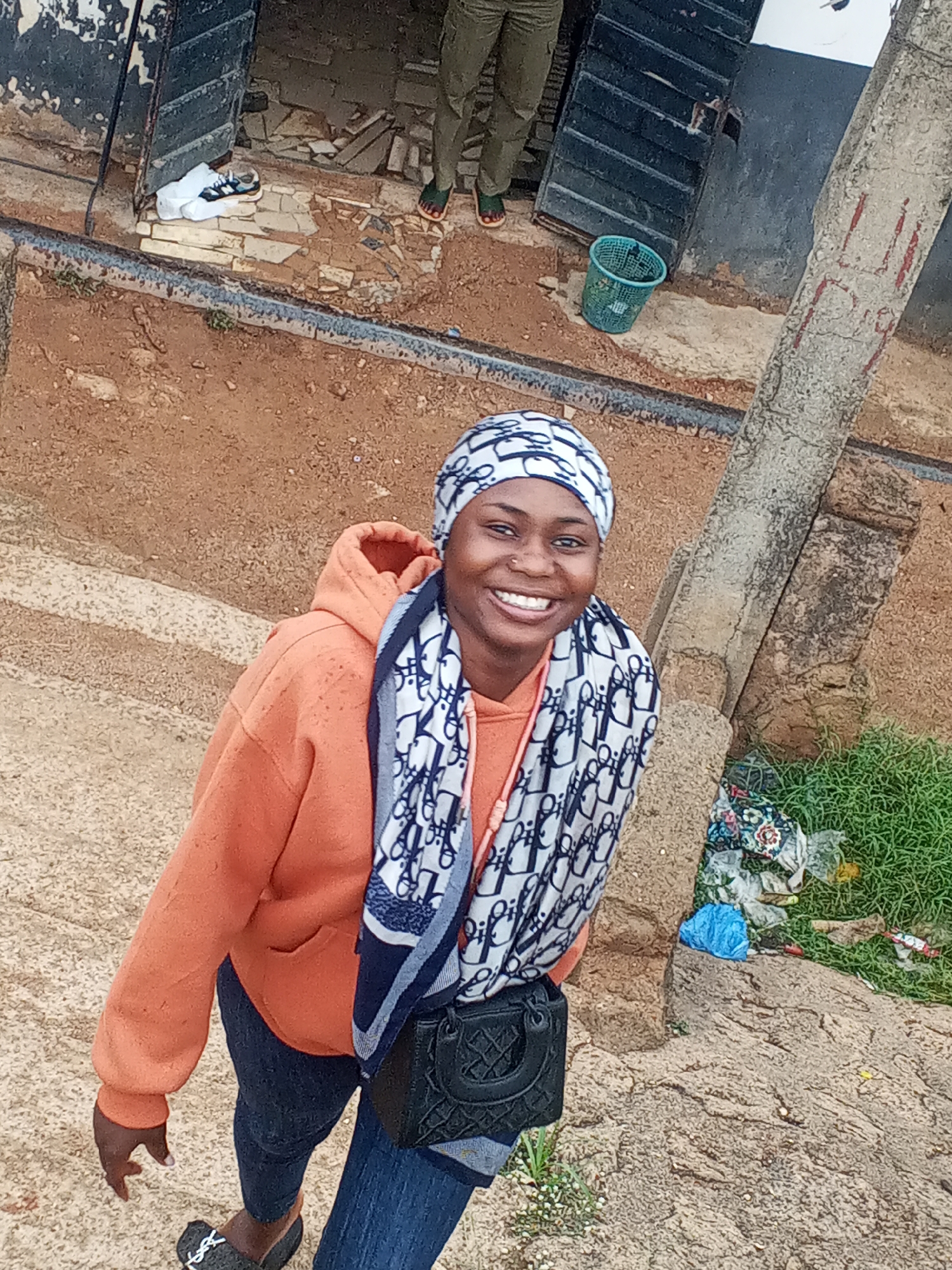 A woman stands on a raised, rocky, and grassy embankment near a building. She is wearing an orange hoodie, jeans, and slides. She has a patterned headscarf and scarf. A small, black purse is slung over her shoulder. A building, trash, and a green basket are visible in the background. A person wearing tan pants and green shoes stands near the building. The location is Gwong, Nigeria.
