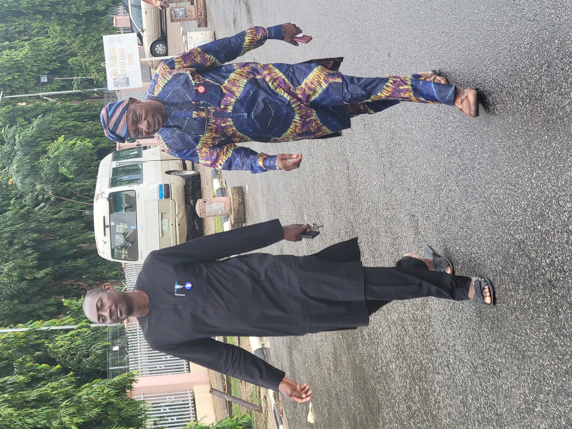 Two men stand outside in a paved area of Abuja, Nigeria, alongside a vehicle and a "No Parking Tow Away Zone" sign.
One man wears a blue and purple patterned top and matching pants with a striped hat. The other man wears a plain black tunic and pants. Both men wear sandals and are facing the camera.
The location appears to be a parking area near a building or office in the city.