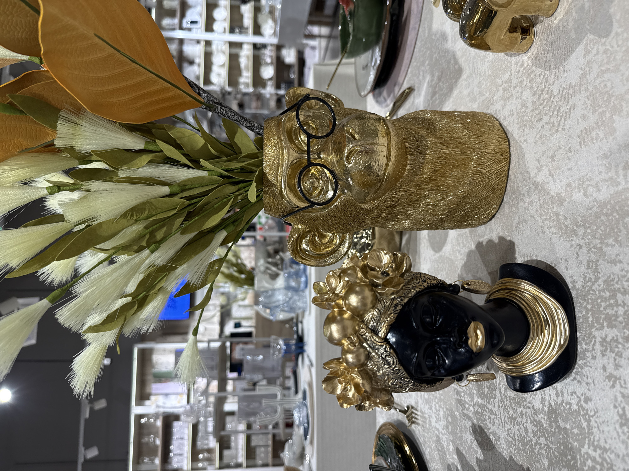 This is a close-up, eye-level shot of decorative items on a table, likely in a retail store. The primary subjects are two gold-colored decorative pieces: a bust of a Black woman wearing an elaborate gold headdress, gold earrings, and gold necklaces, and a monkey-shaped vase with round glasses. The monkey vase holds a floral arrangement of white, feathery plumes and green leaves, with a large, orange leaf extending out. To the right of the monkey vase, there are two small, gold elephant-shaped salt and pepper shakers. In the background, shelves filled with various dishware and glassware are visible, suggesting a home goods or department store. The lighting is bright, and the overall scene is well-lit. There are no people visible in the image. The location is not identifiable within the city of Izhevsk, Russia based on the visual information provided.