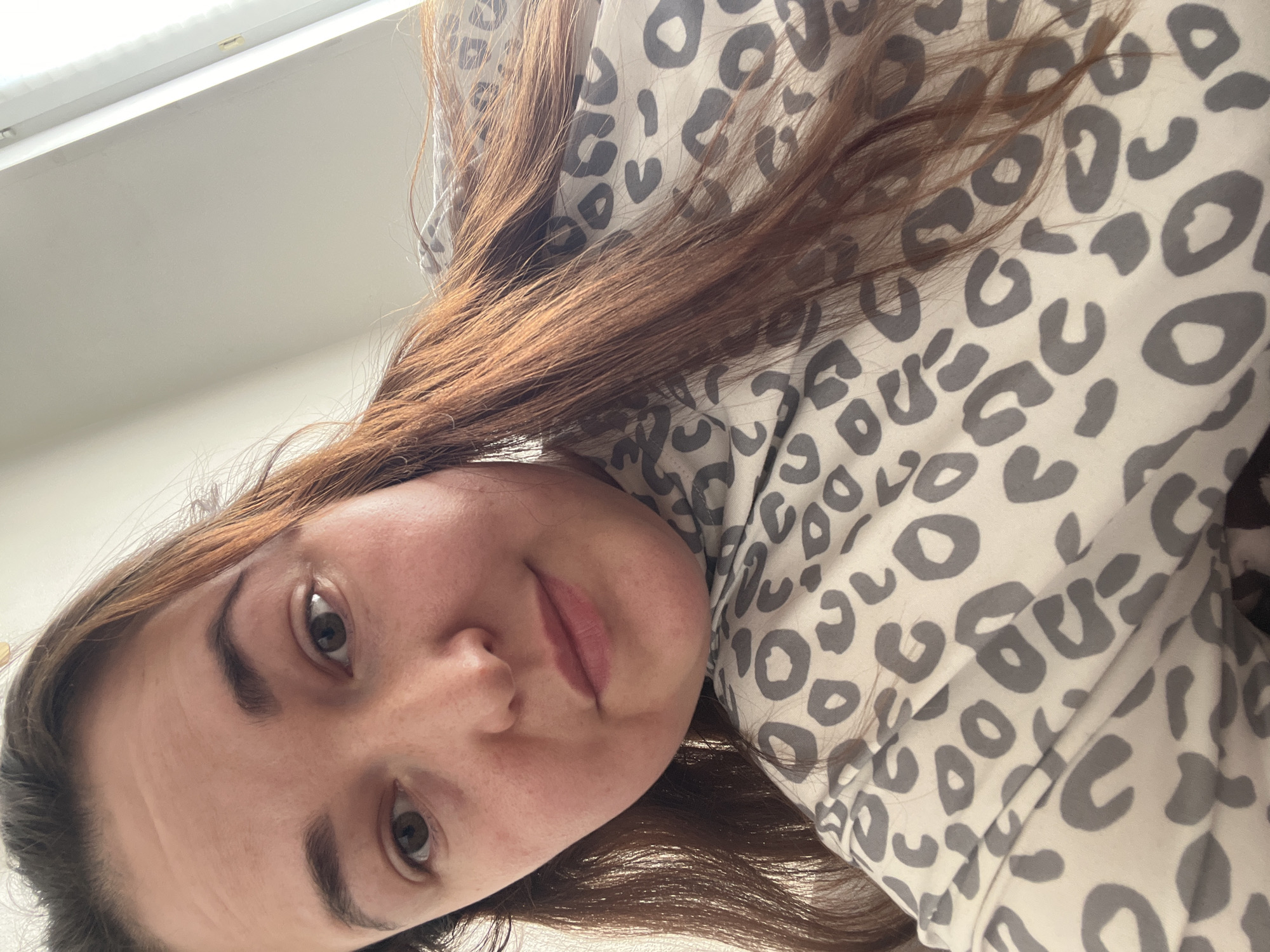 A woman with long, reddish-brown hair is looking down at the camera in a selfie. She is wearing a light-colored, long-sleeved shirt with a grey leopard print. The background appears to be a lightly colored wall with natural light coming from the right side of the frame. The image is taken from a low angle. No specific location within Chelyabinsk, Russia can be determined from the provided image.