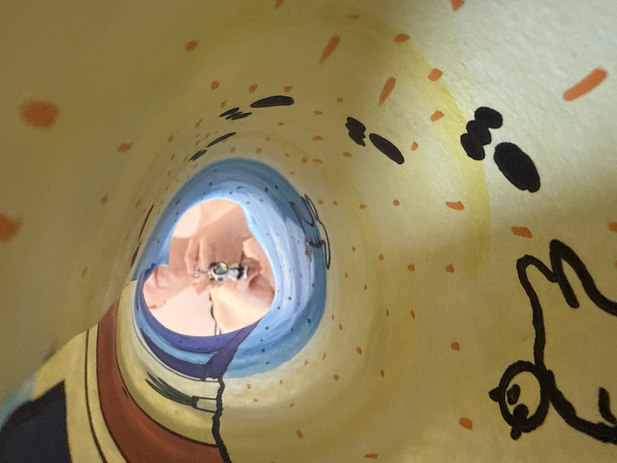 The image shows the interior of a children's drawing, possibly a tunnel or tube, painted on a yellow background with scattered orange and black markings. At the far end of the tunnel, there is a view of a hand wearing a ring or other decorative object. The tunnel is painted with blue and other colored stripes. The style of the drawing is cartoonish. There is no information indicating the location is in the city of Izhevsk, Russia.