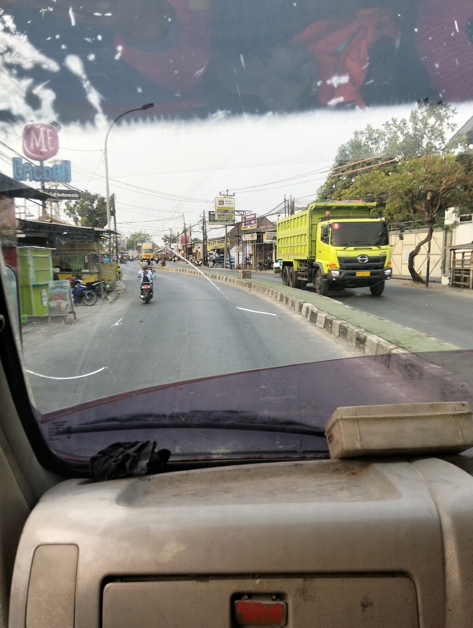 The image is taken from inside a vehicle, looking out through the windshield. The perspective suggests the vehicle is driving on a road. In the foreground, the dashboard of the vehicle is visible, with a dark cloth or rag placed on it.
The road in the image appears to be a main thoroughfare, with multiple lanes of traffic. On the right side of the road, there is a prominent bright yellow dump truck. The truck has a Hino logo on its grille. The truck is moving in the same direction as the vehicle from which the photo is taken.
On the left side of the road, there are several buildings and stalls, possibly shops or food vendors, with various signs in Indonesian. One sign prominently displays "ME Griban". There are parked motorcycles on the left side. Further down the road, other vehicles and motorcycles are visible, indicating moderate traffic. The sky is overcast.
The image is likely taken in a commercial or semi-urban area of Semarang, Indonesia, given the signage and the type of businesses visible. The presence of a dump truck suggests it could be an area with construction or industrial activity, or simply a route for heavy vehicles. The overall scene depicts a typical day on a busy road in Indonesia.
